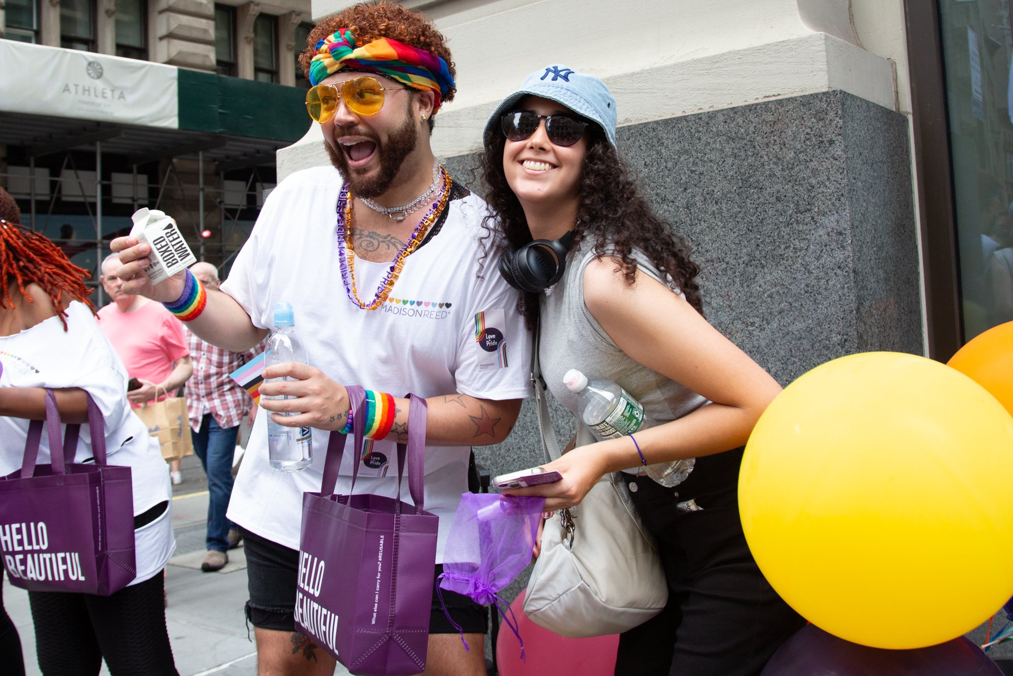 People celebrating at a Pride event with rainbow accessories, reusable shopping bags, water bottles, and colorful balloons.