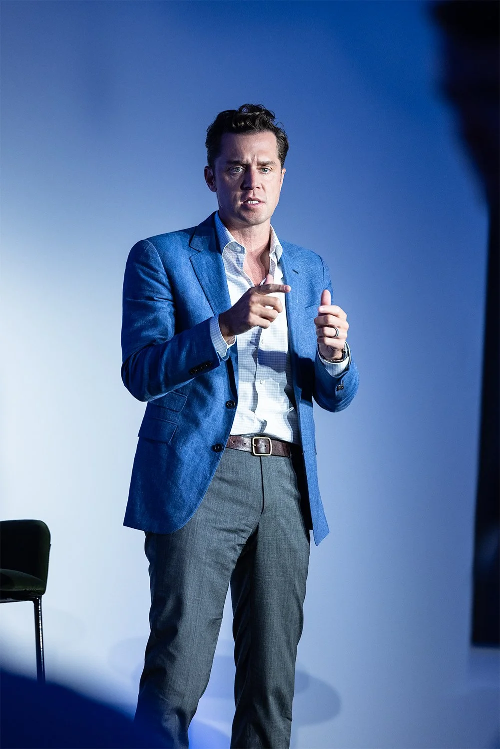 A man in a light blue suit jacket and gray trousers giving a presentation or speech, pointing with his right hand, against a plain gray background.