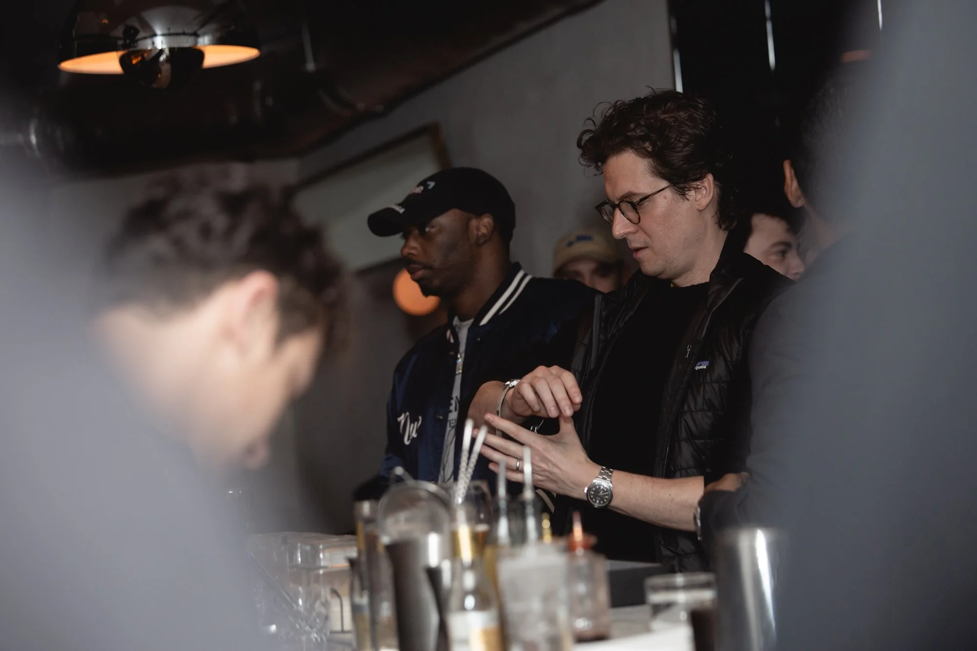 A group of people are gathered around a table or bar, engaged in conversation, with drinks and bottles in the foreground.