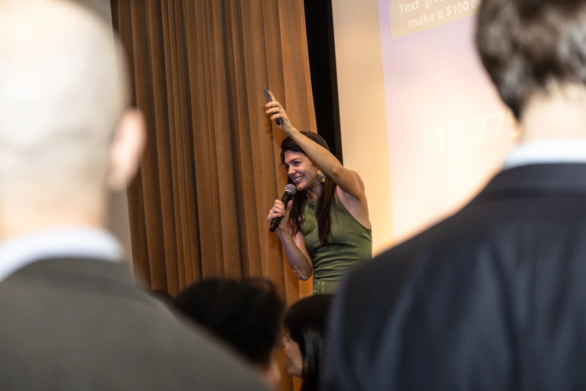 A woman smiling and holding a microphone, raising her right arm, standing in front of a projector screen amid an audience.