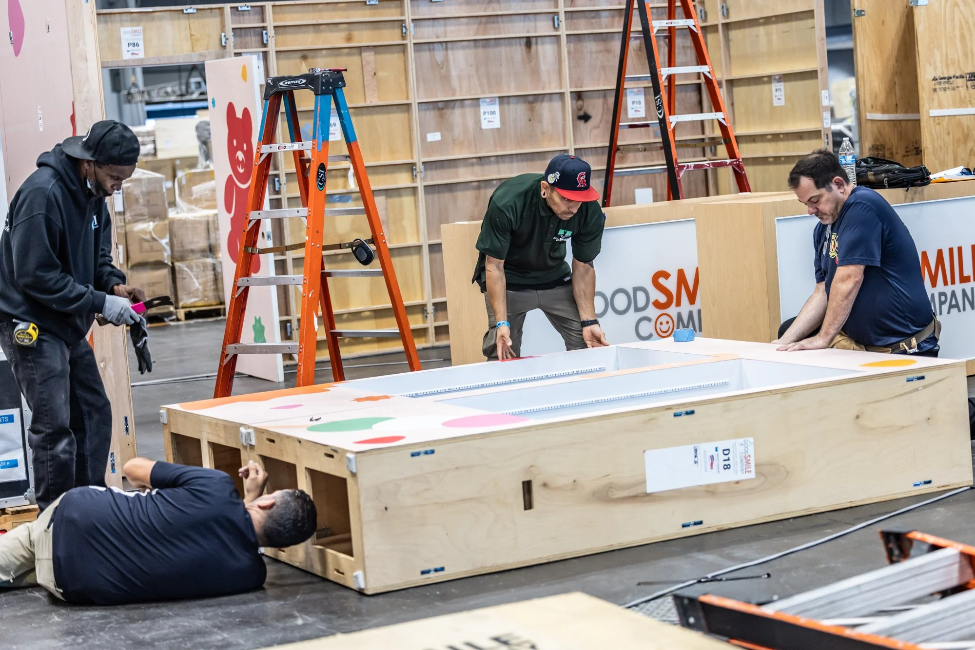Four men assemble a large wooden display booth inside an event venue, with ladders, crates, and signage visible.