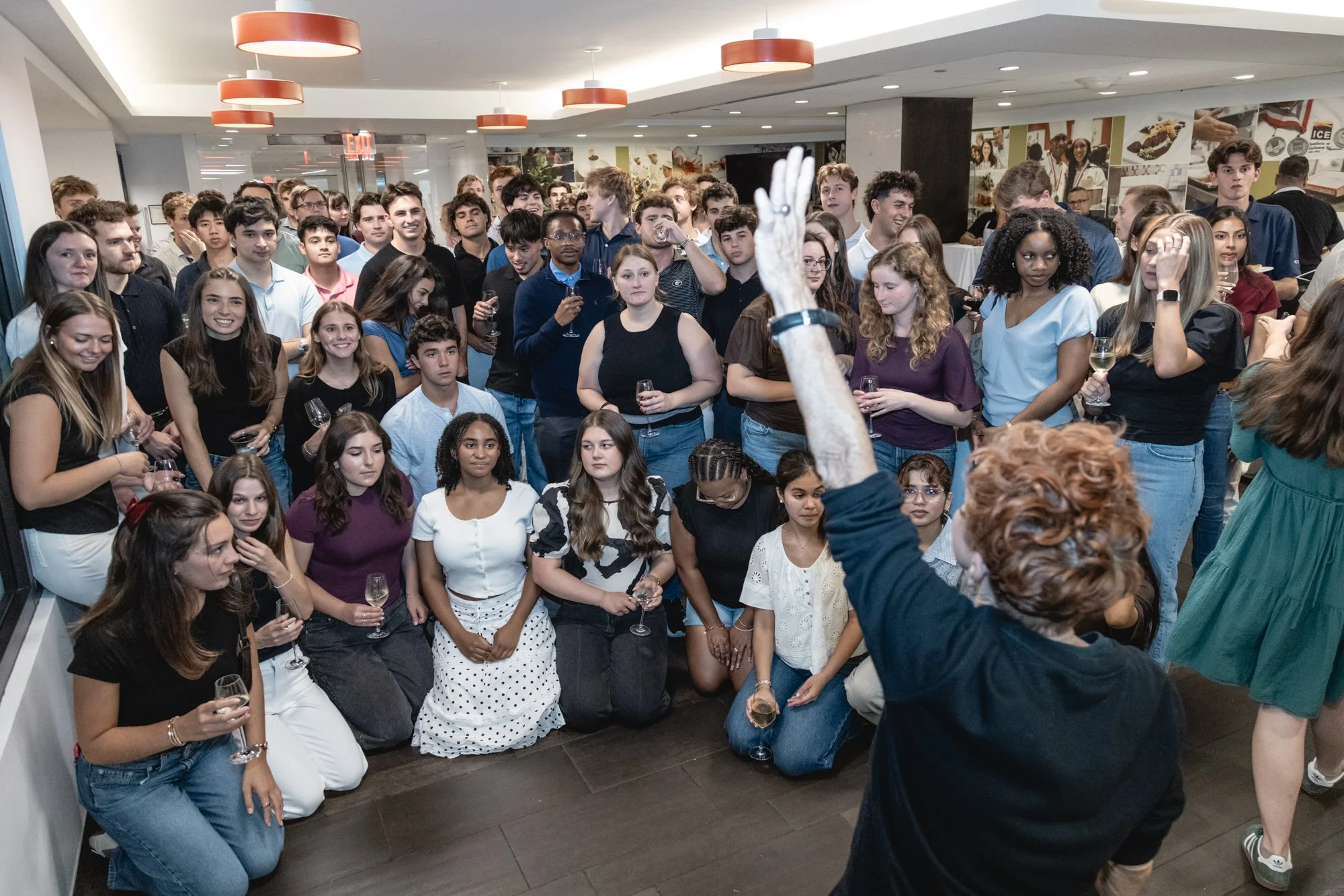 Group of young adults at an indoor event, some standing and some kneeling, holding glasses of wine and listening to a speaker with their hand raised.