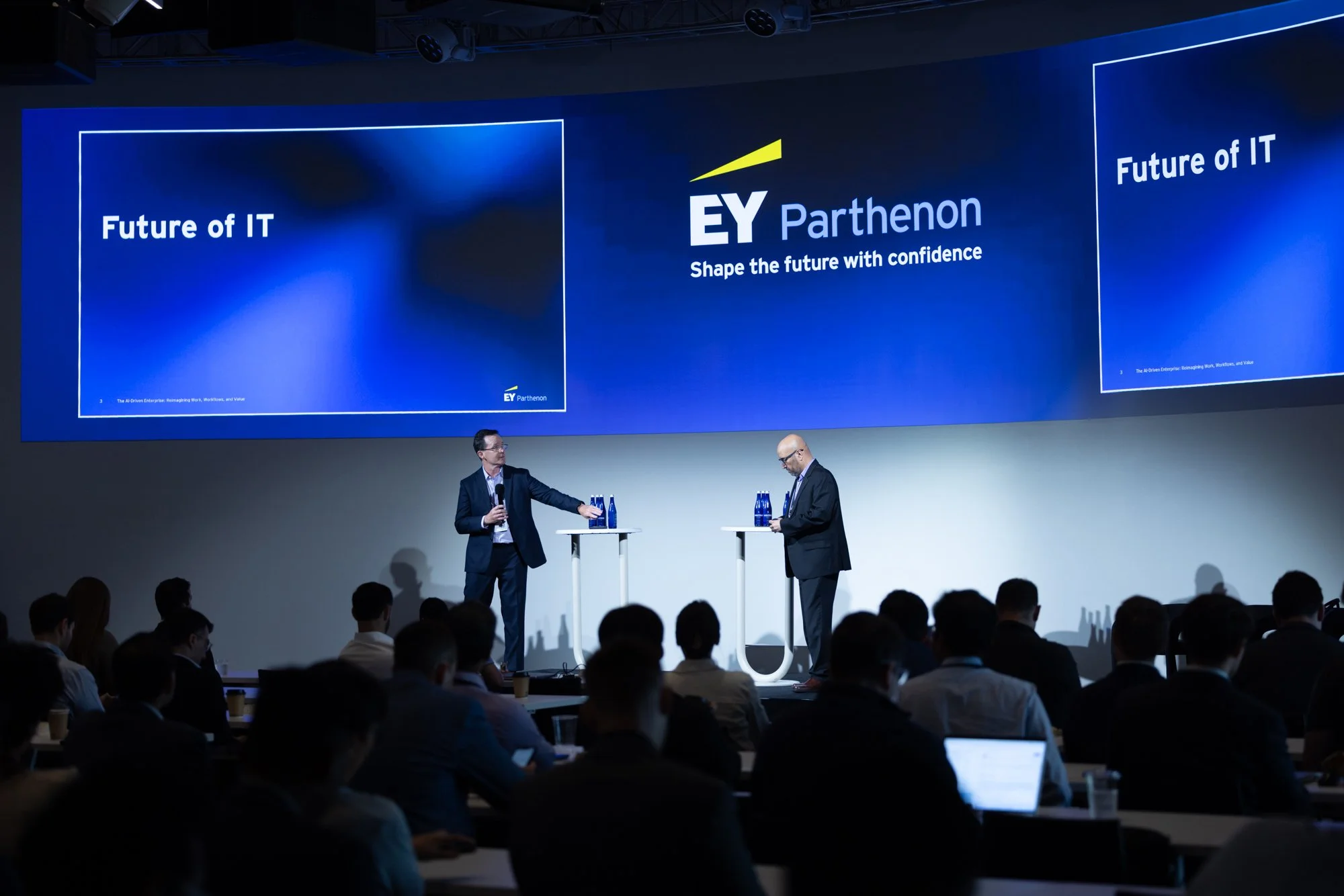 A large conference room with audience members sitting and watching two men on stage. The stage has two tall tables with water bottles. Behind the men, a large screen displays the EY Parthenon logo, the phrase 'Future of IT,' and the tagline 'Shape th