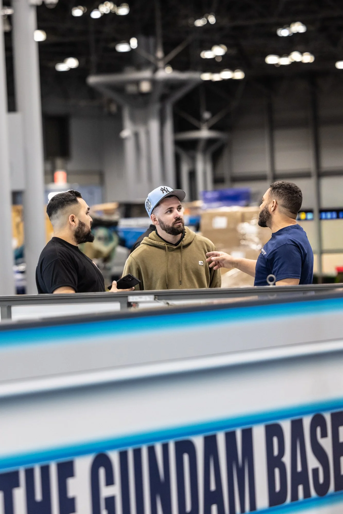 Three men talk beside a New York Yankees batting cage in an indoor baseball training facility.