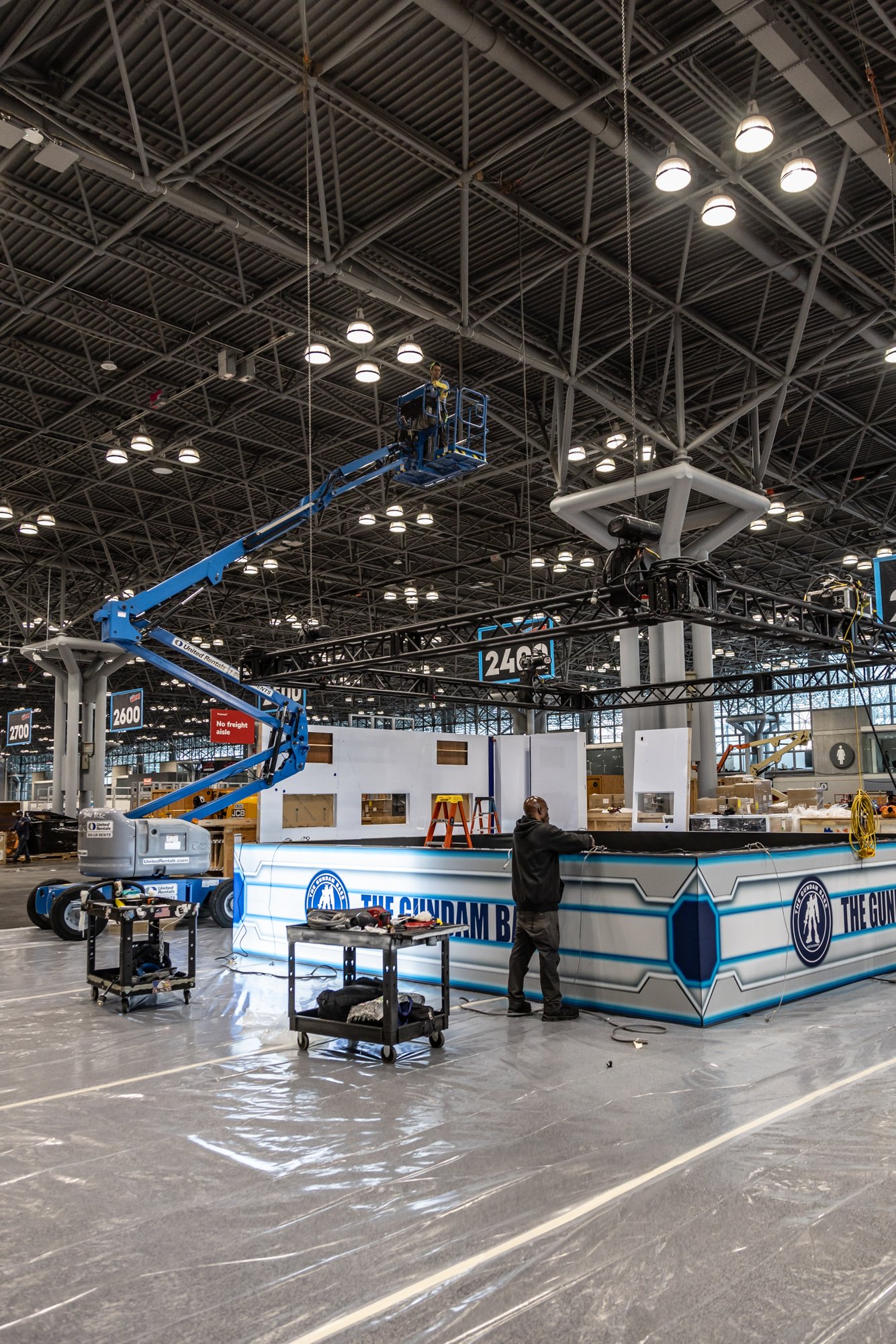Worker assembles a branded booth inside a large convention center using a blue boom lift.