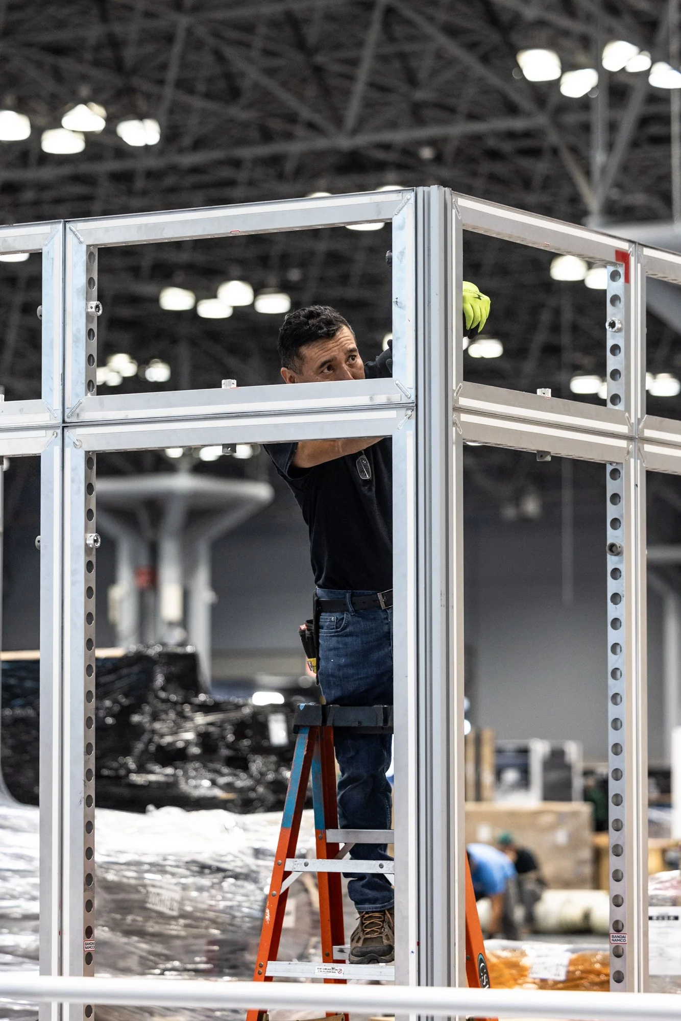 Worker in a safety vest assembling a metal booth structure from an orange step ladder inside a convention center
