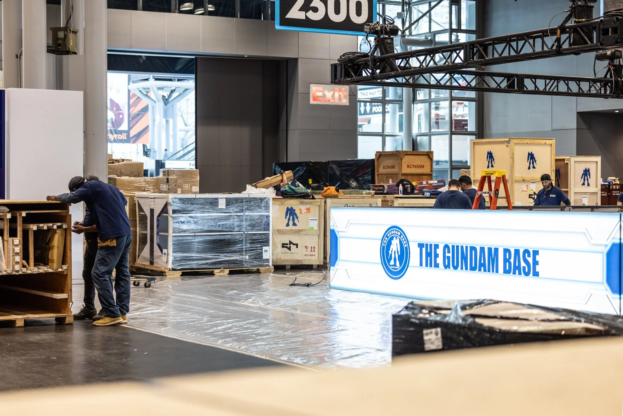 Workers set up The Gundam Base display at an indoor convention venue surrounded by shipping crates, pallets, lighting equipment, and scaffolding.