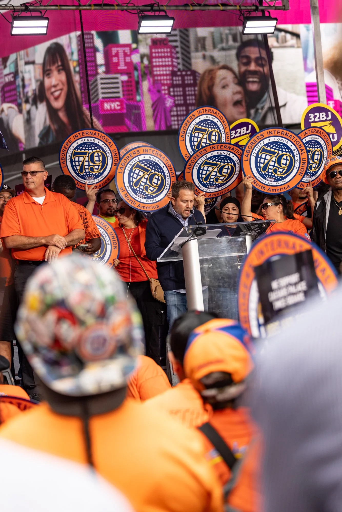 A large crowd gathered at a rally or demonstration, with people holding circular signs that say '79' and 'Labourers' International Union of North America' with a globe logo. There are people in orange shirts, some at a podium speaking into a micropho