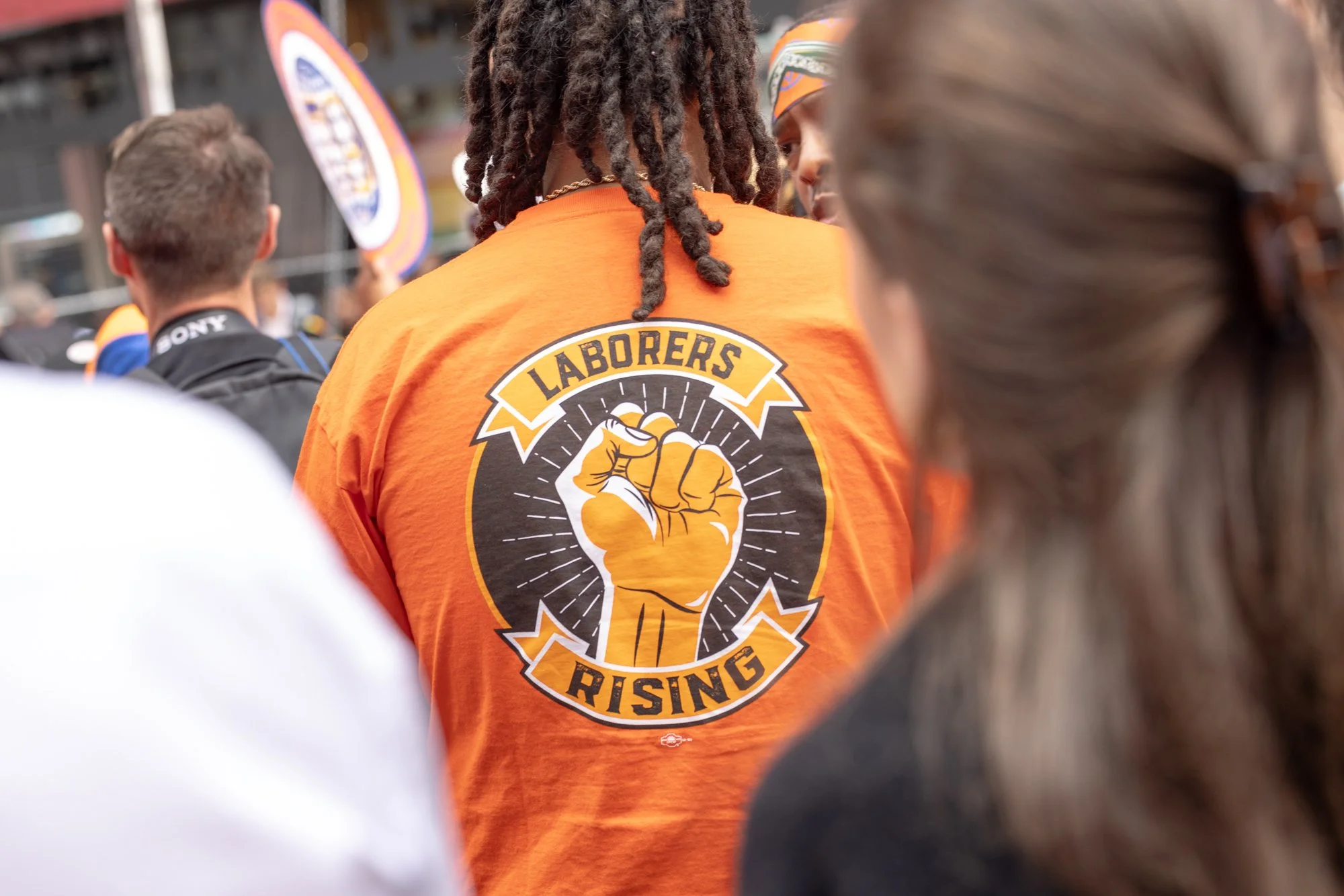 Back view of a person with dreadlocks wearing an orange shirt with a raised fist logo and text that reads "Laborers Rising" at a protest or rally.