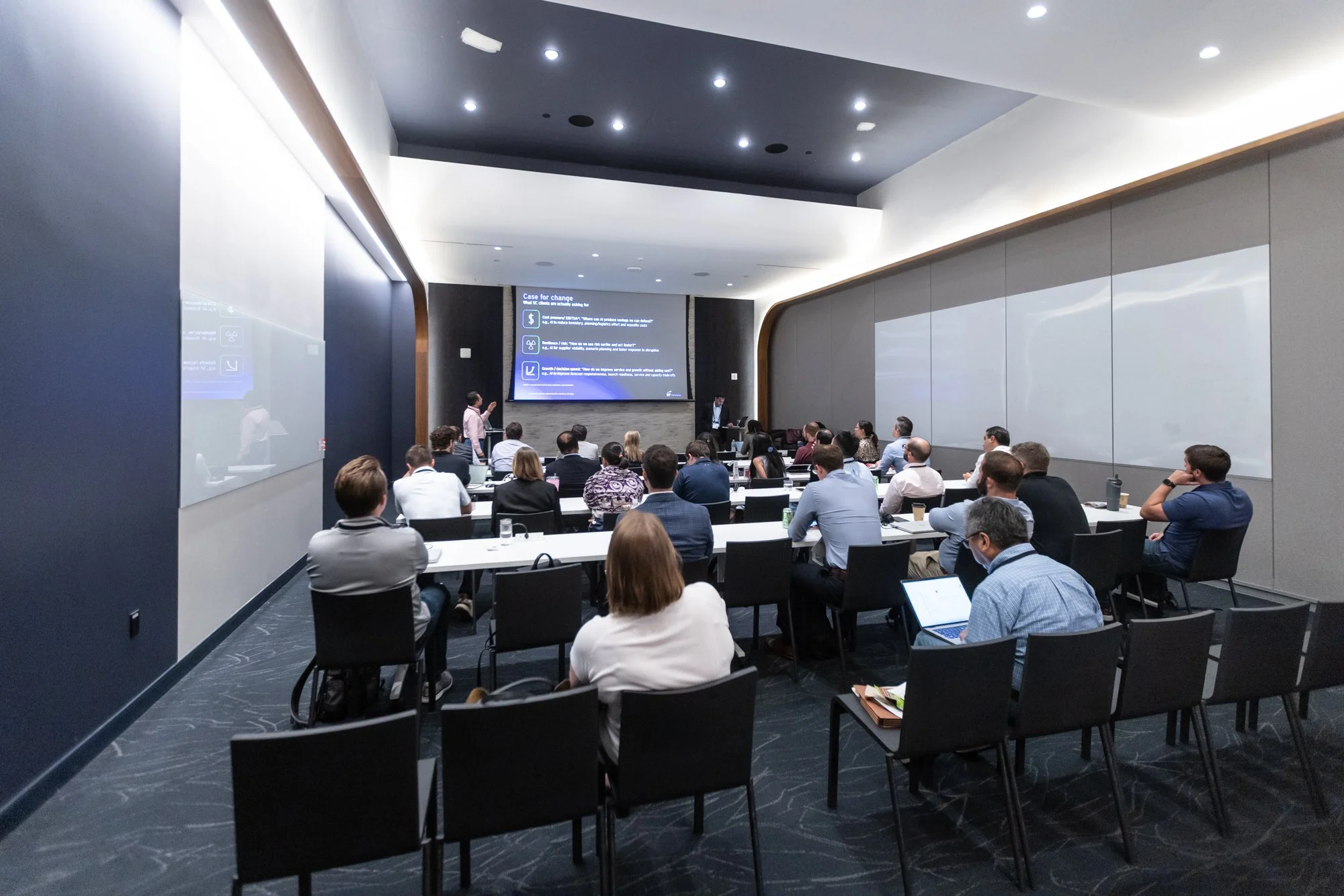 A conference room filled with seated attendees facing a stage with a large screen showing a presentation. A speaker stands at the front of the room near a podium, with another person operating a laptop. The room has modern decor with curved archways 