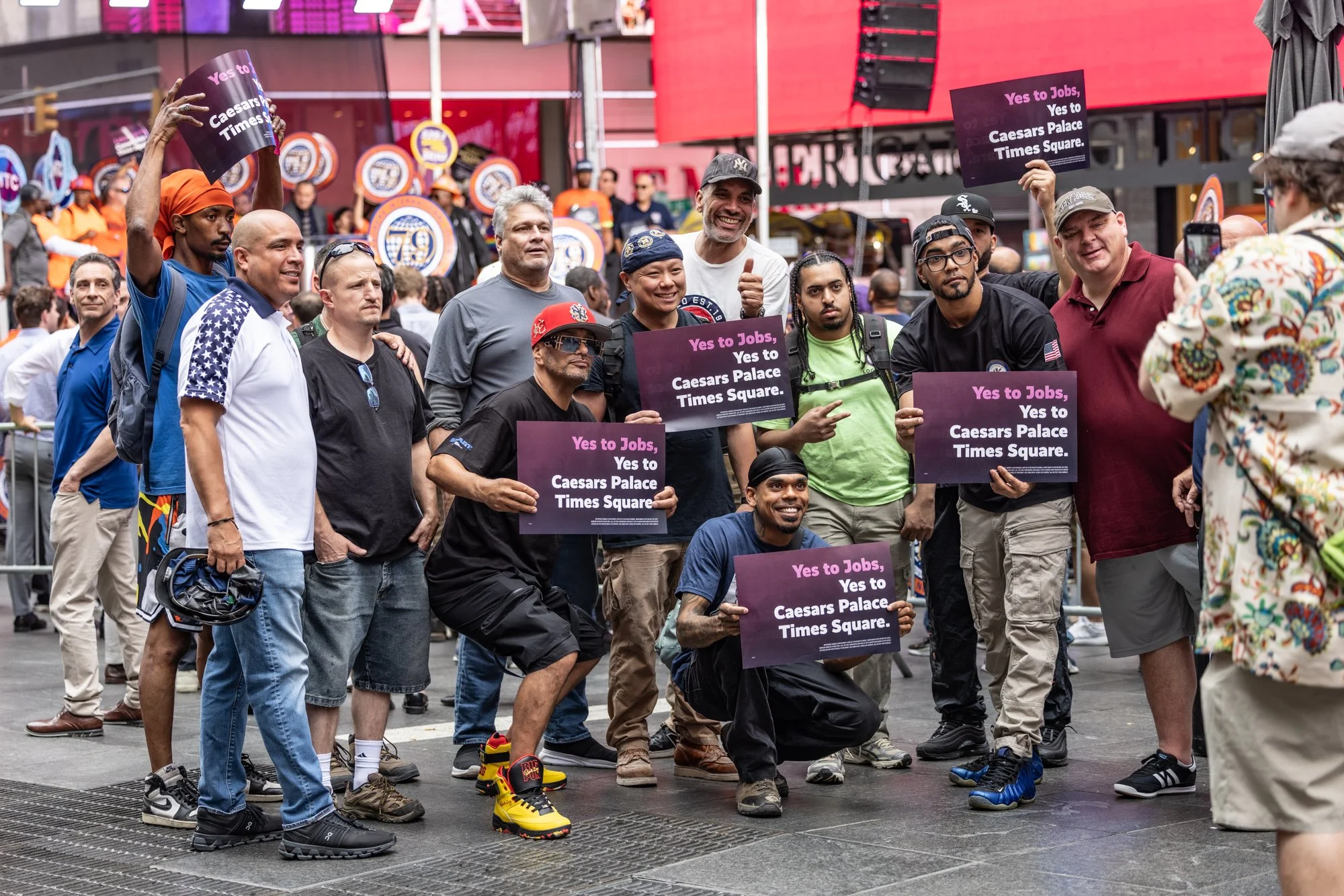A diverse group of people, including men and women of different ages and ethnicities, participating in a protest or rally in Times Square. Several individuals are holding purple signs that read 'Yes to Jobs, Yes to Caesars Palace Times Square.' Some 