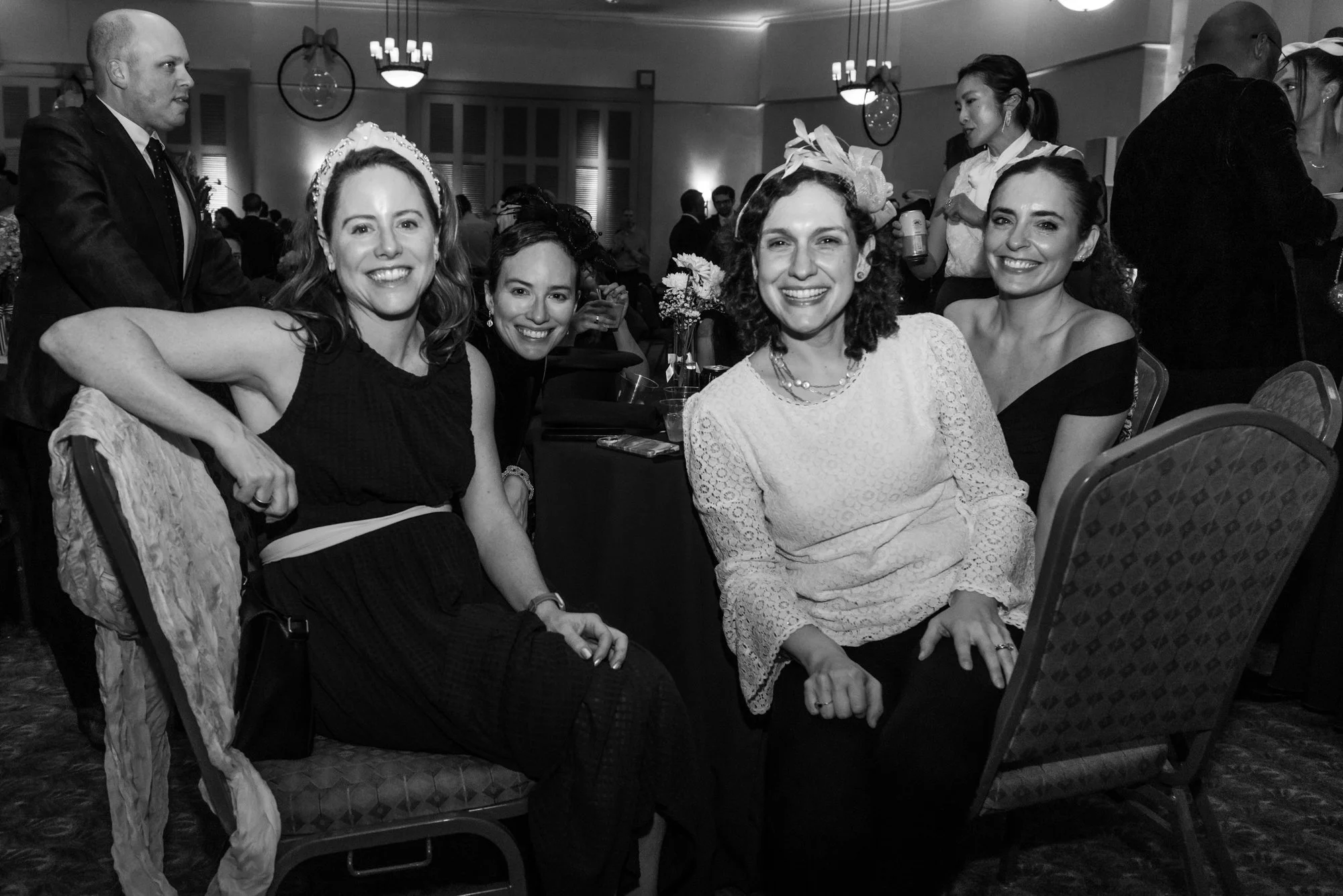 Young women sitting at a banquet table, dressed elegantly, smiling at the camera, with some wearing fascinators and jewelry, at a formal event or celebration in a decorated hall.