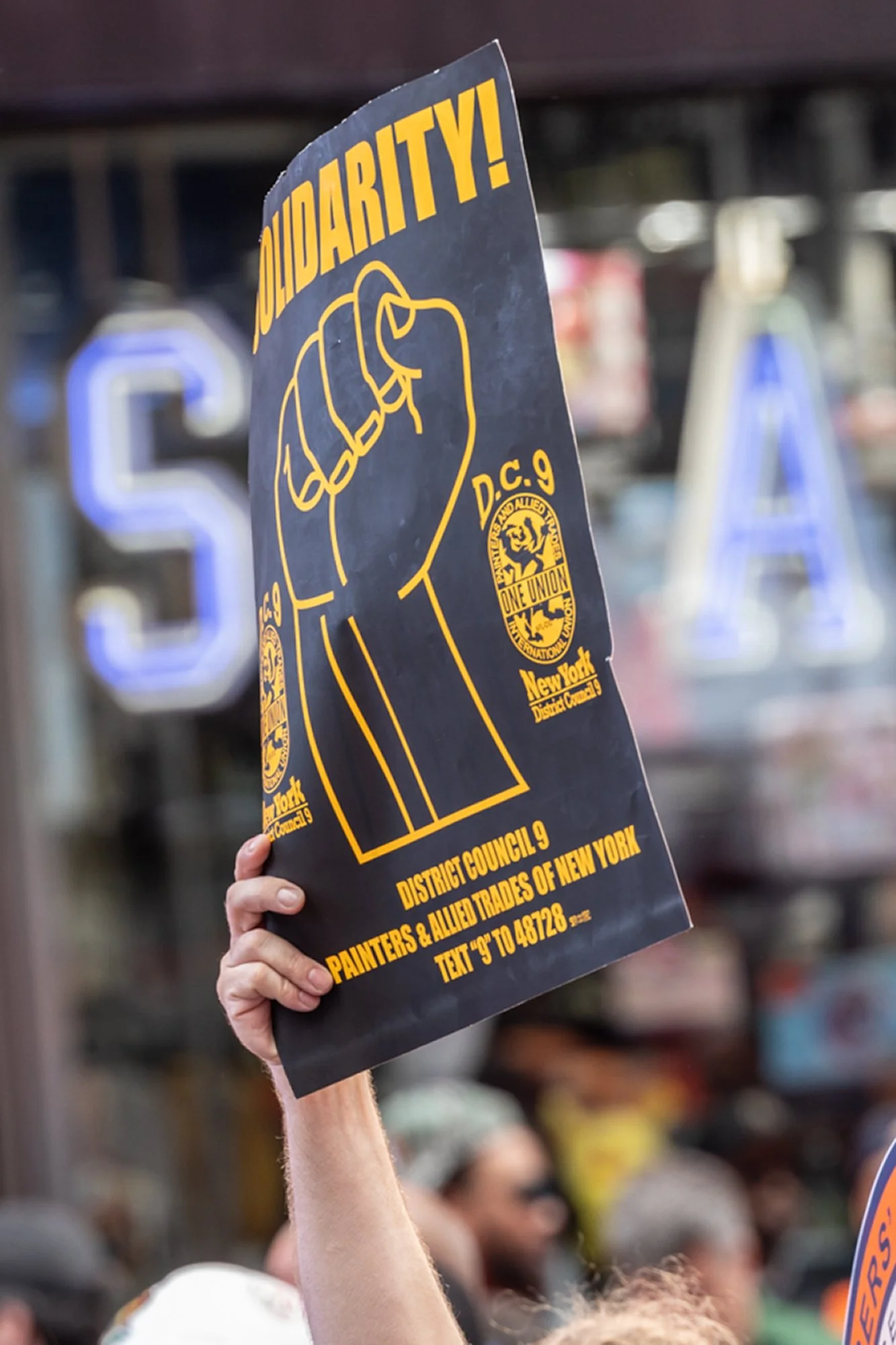 A protestor holds a sign with the words "Solidarity!" and a clenched fist illustration, during a public demonstration with a crowd and blurred background of store shelves.