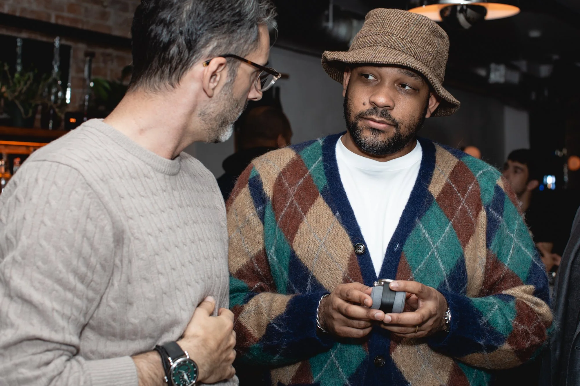 Two men engaged in conversation at an indoor event. One man, with dark hair, glasses, and a beige sweater, is looking at another man, who is wearing a brown bucket hat, a colorful argyle cardigan, and a white T-shirt, holding a smartphone. In the bac