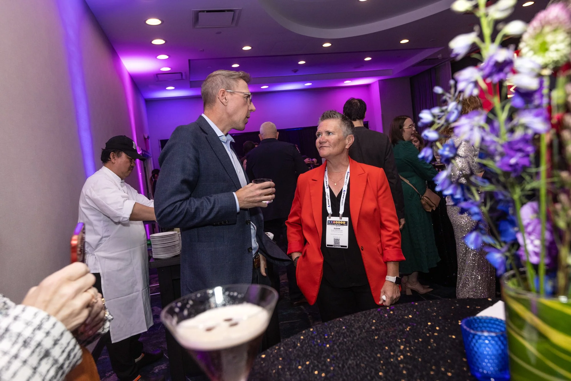Two people are engaged in conversation at a social event. The man on the left, with short gray hair, wearing glasses and a dark blue blazer, holds a glass. The woman on the right, with short gray hair, wearing a bright orange blazer and a conference 