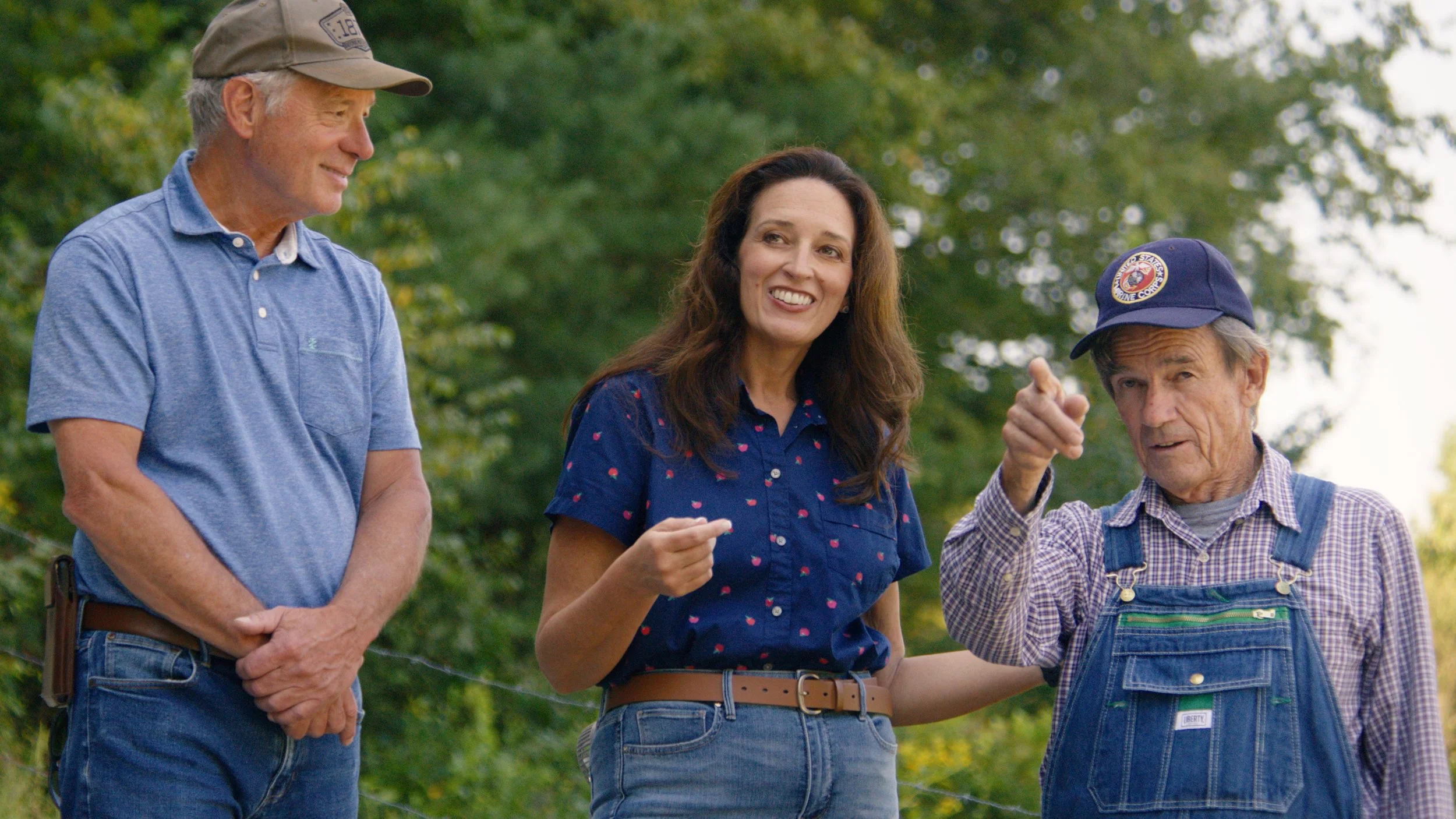 Three people outdoors, an elderly man wearing a hat and overalls gesturing, a woman with long brown hair smiling and gesturing, a middle-aged man in a cap and sunglasses standing nearby with a tree background.