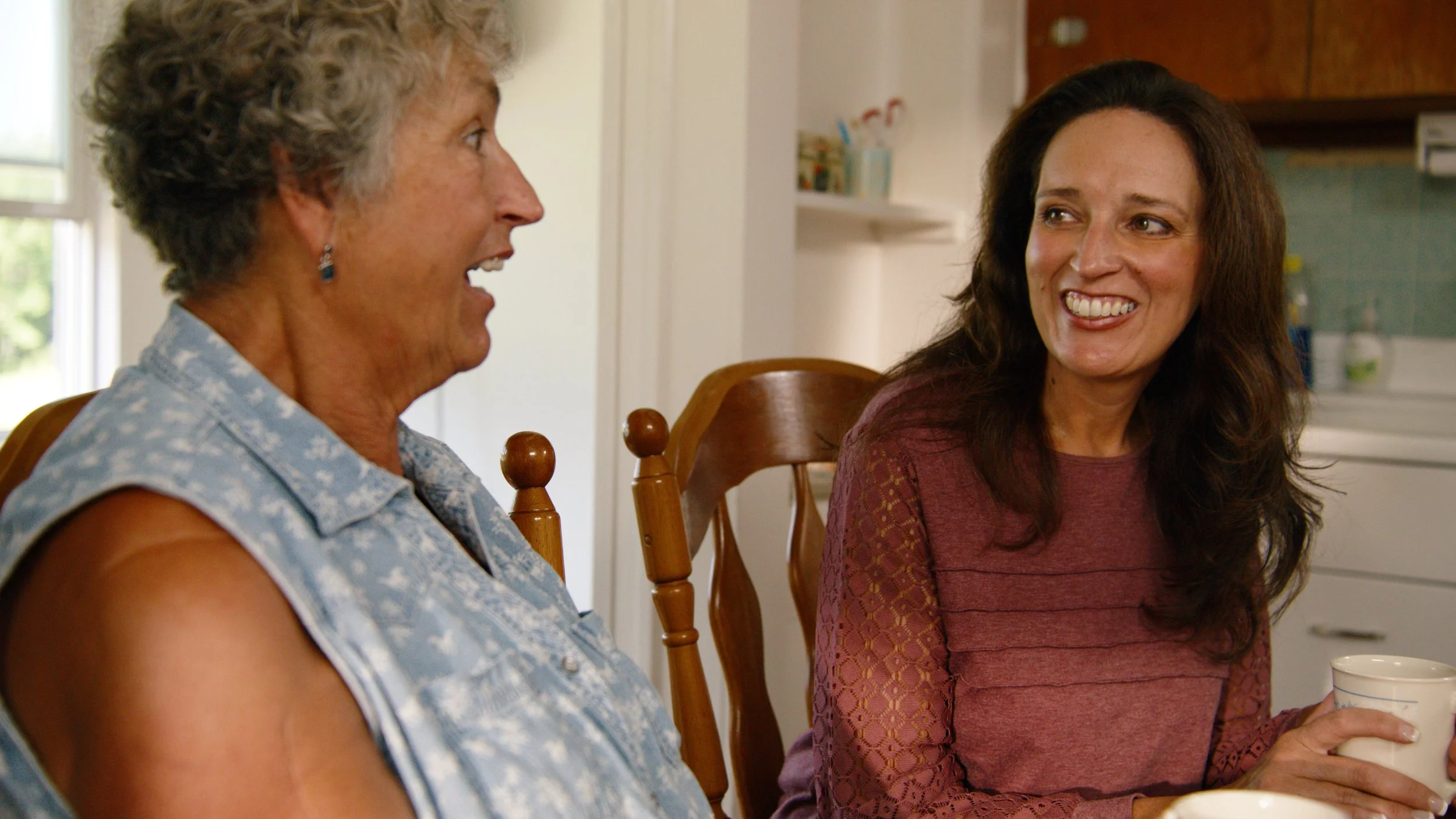Two women sitting at a dining table, smiling and engaging in conversation in a kitchen setting.