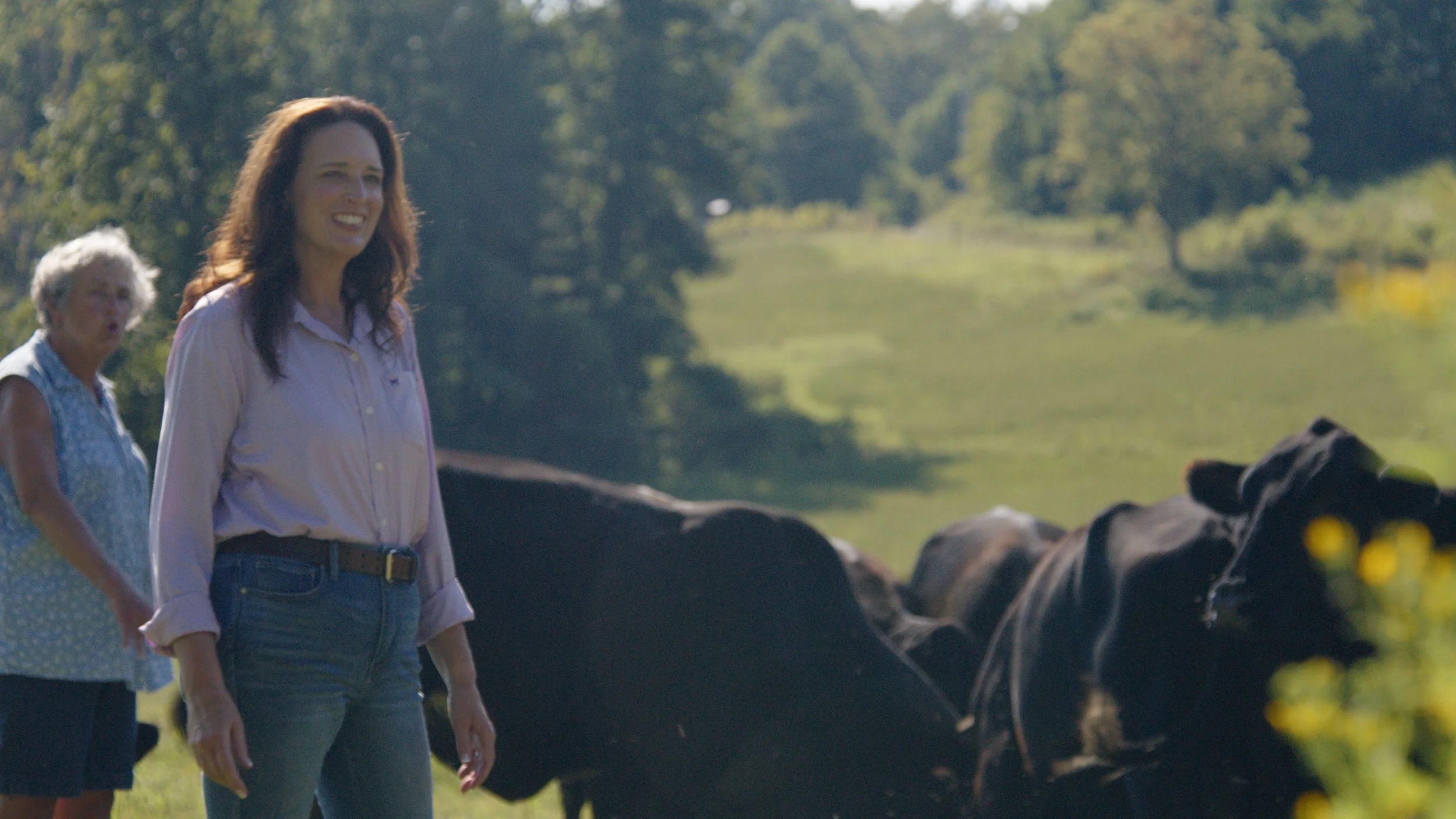Two women walking outdoors in a scenic, green park with trees and rocks, accompanied by horses.