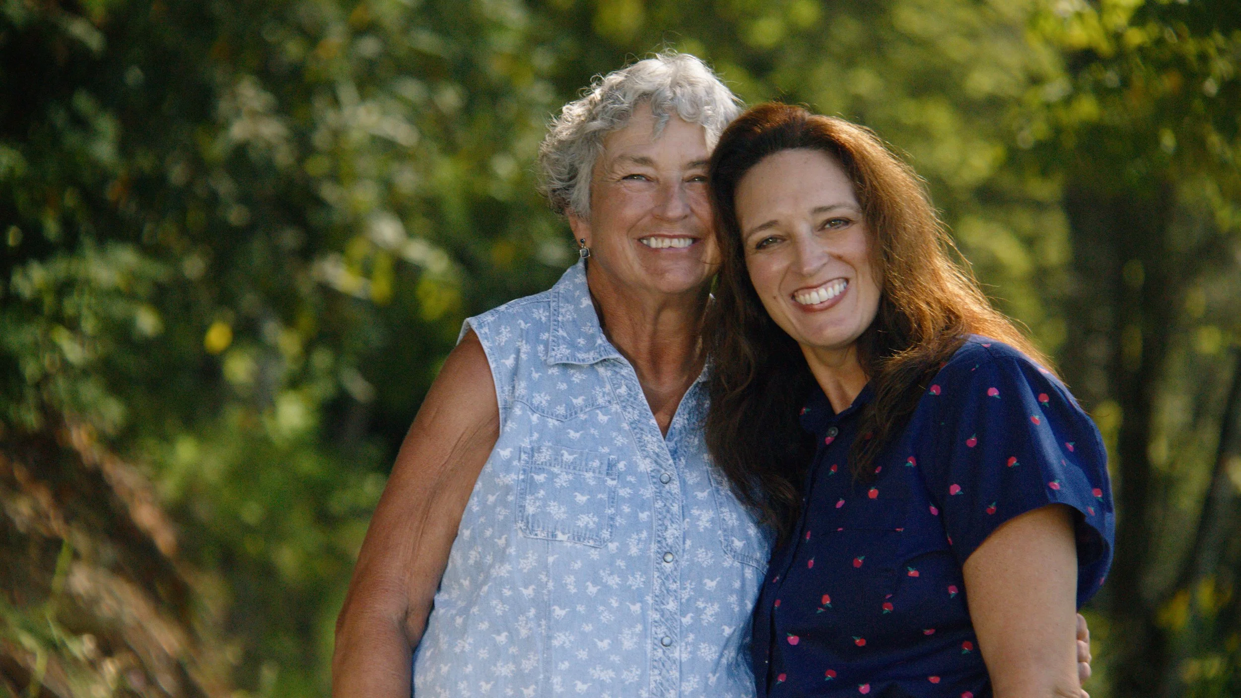 Two women smiling and standing outdoors, close together with greenery in the background.