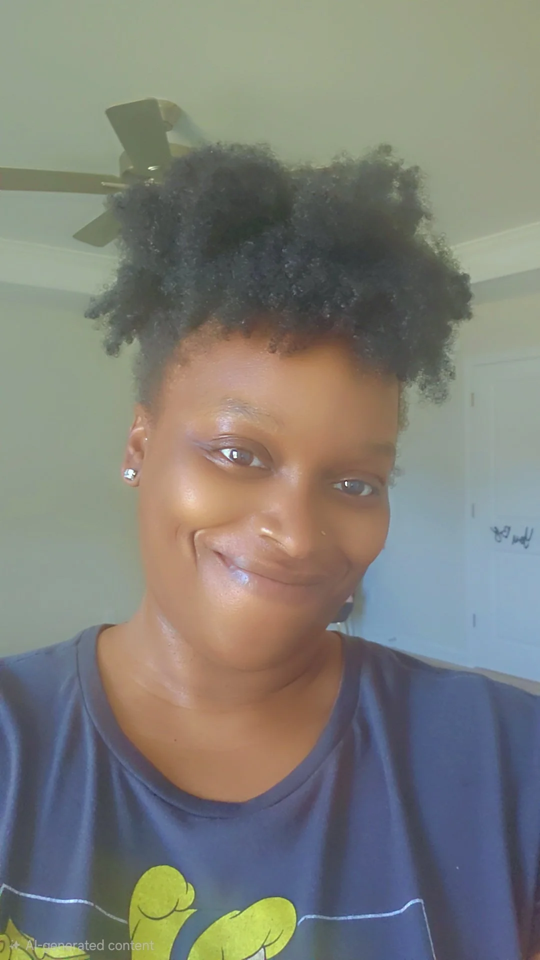 Close-up photo of a smiling woman with natural curly hair, wearing earrings and a dark T-shirt with a yellow graphic. She is indoors with a ceiling fan and a door visible in the background.