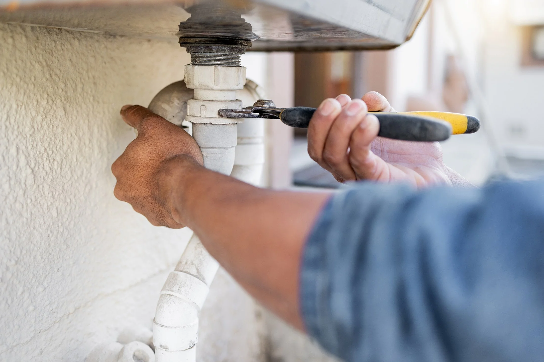 Person using a wrench to tighten a pipe fitting beneath a sink, with multiple white PVC pipes and a textured wall in the background.