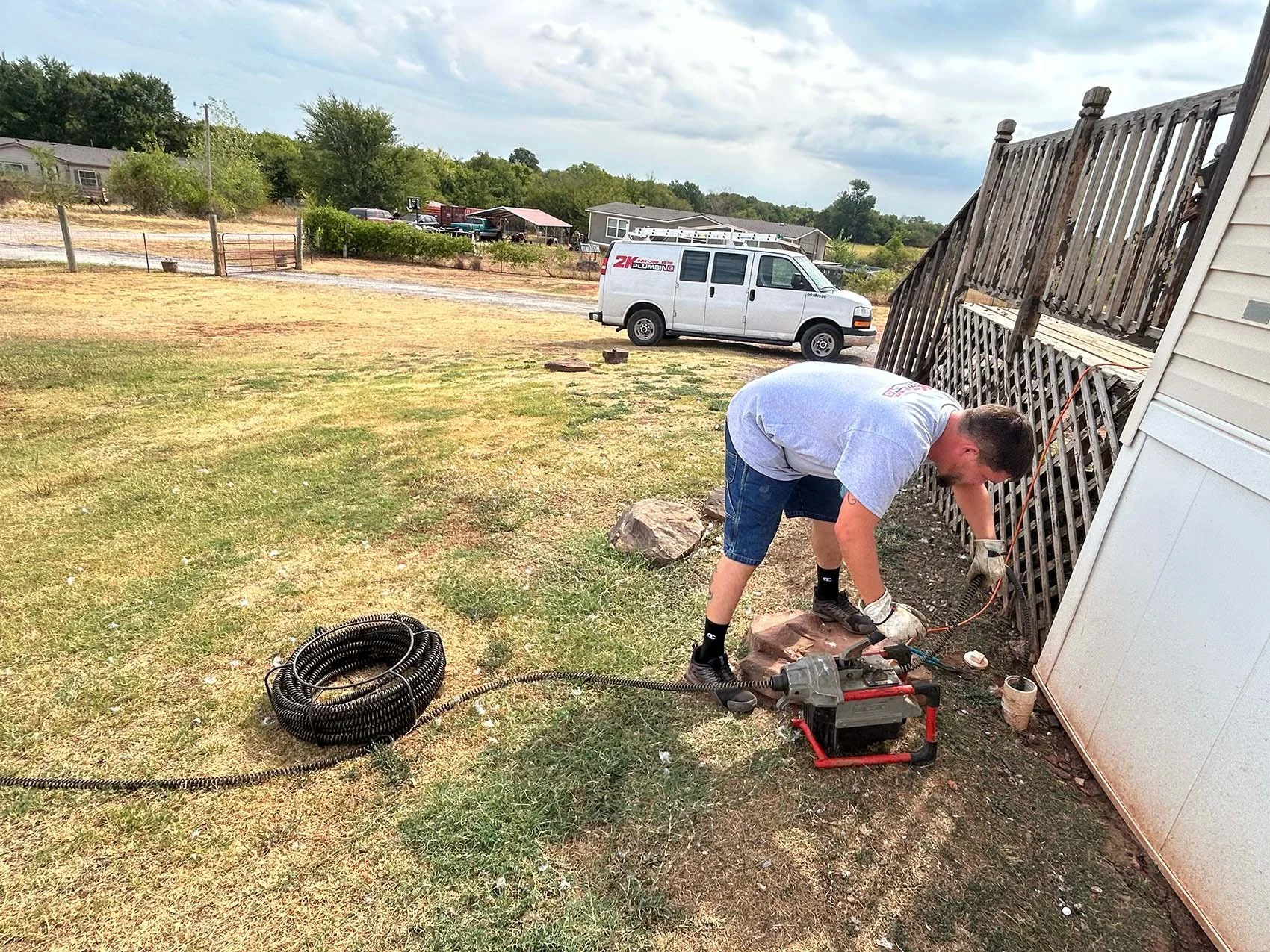 A man working on outdoor electrical wiring near a damaged wooden fence, with a white service van labeled '2K Plumbing' parked on the grass nearby.