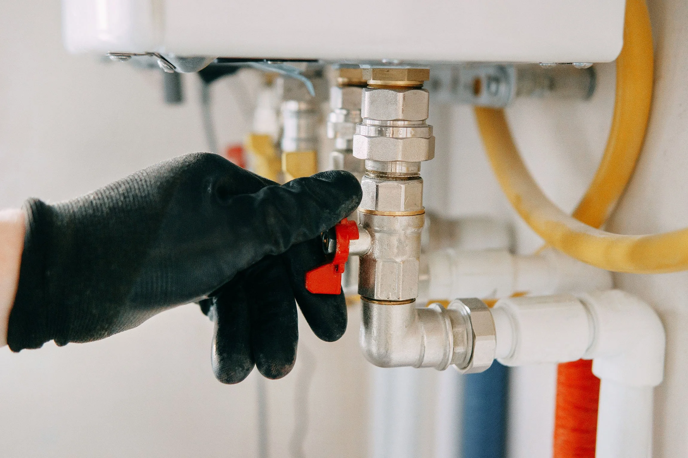 Close-up of a gloved hand adjusting plumbing pipes beneath a fixture, with various metallic fittings and yellow hoses visible.