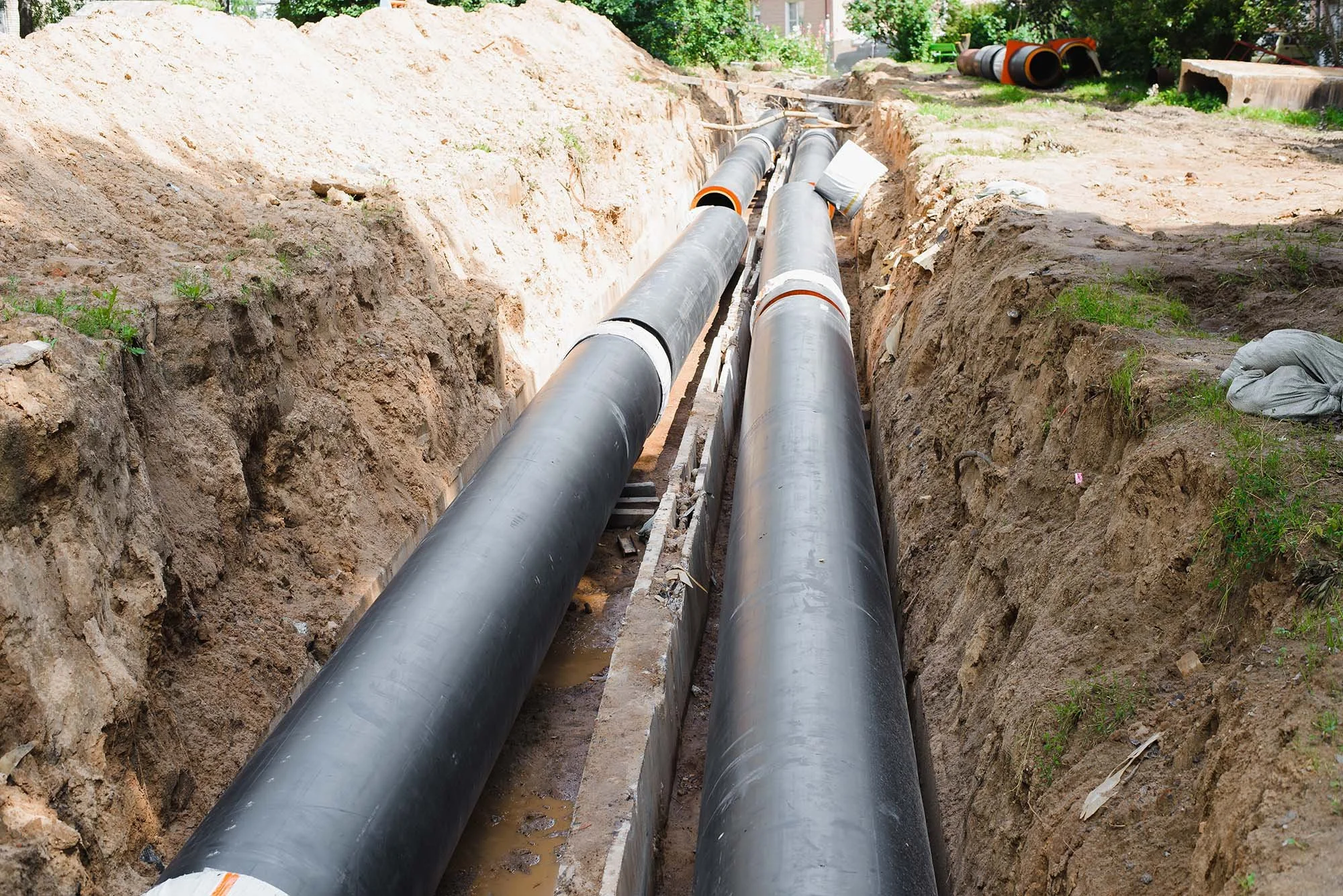 Construction site with two large black pipes in a trench, surrounded by dirt and soil, with some greenery and trees in the background.