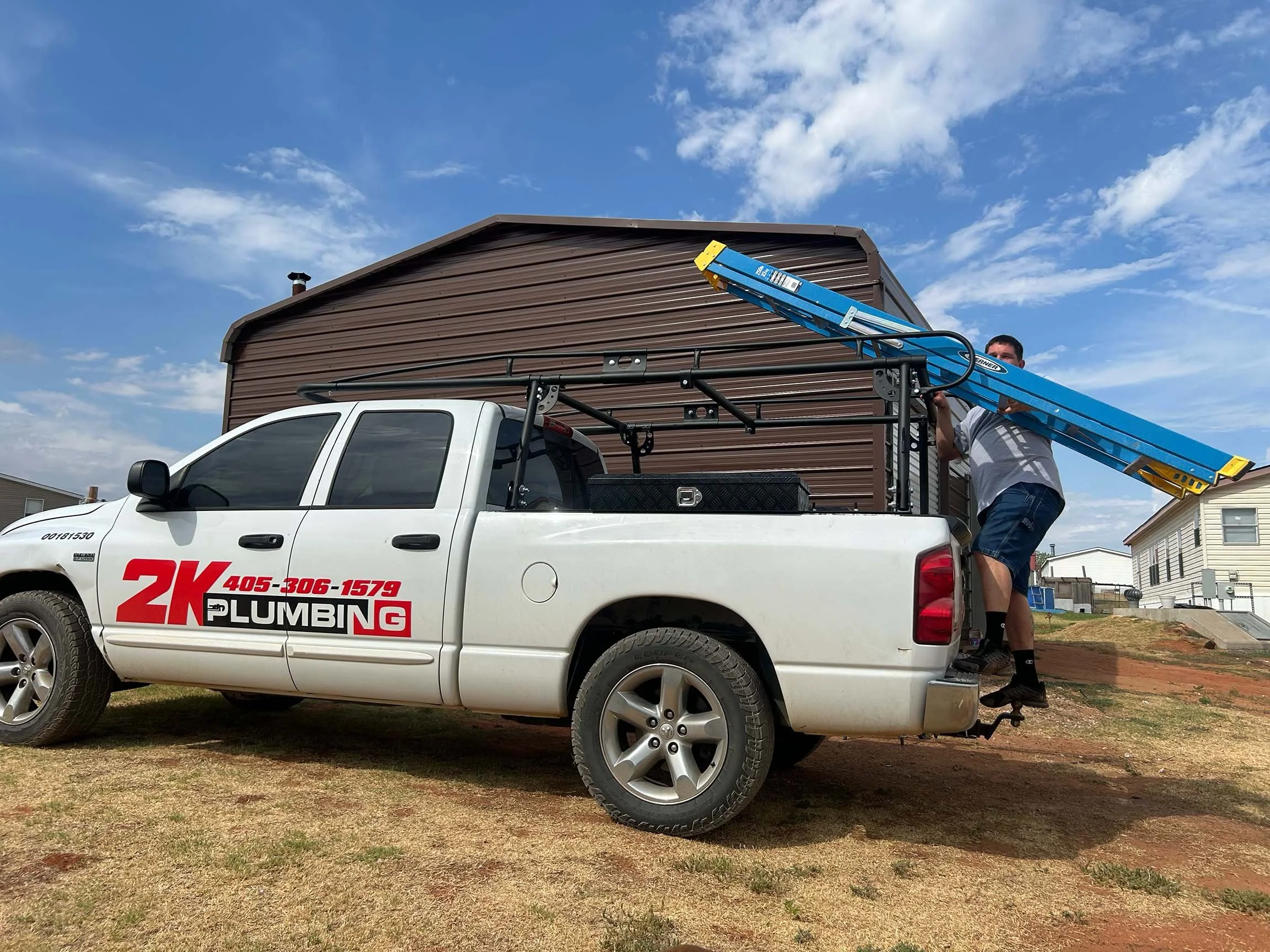 A man standing on the truck's bumper, holding a blue ladder and working on the side of a brown building in a residential area under a partly cloudy sky.