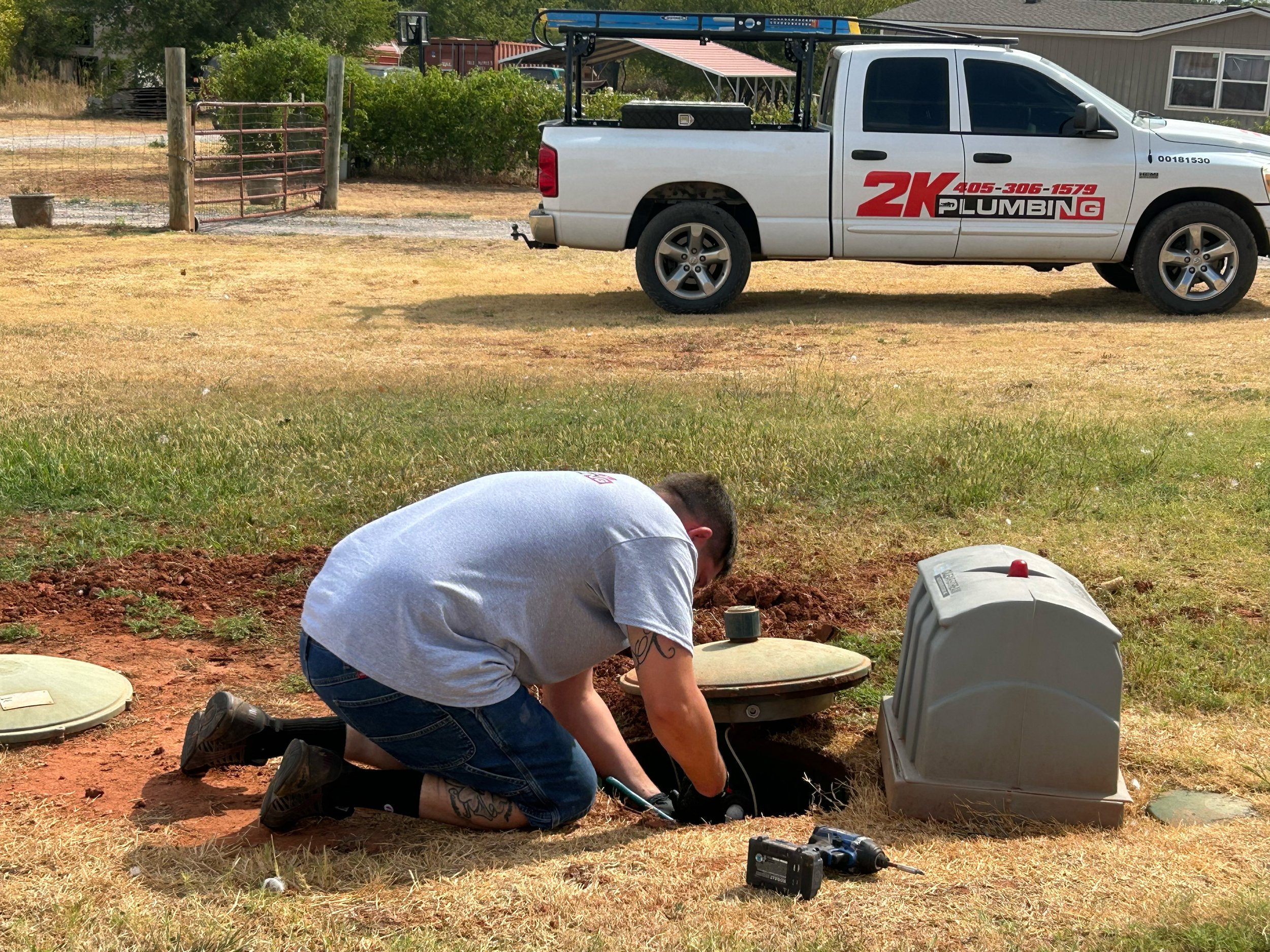 A person working on a plumbing issue underground next to a septic tank or drain system, with a work vehicle parked nearby in a yard with dry grass and a residential building in the background.