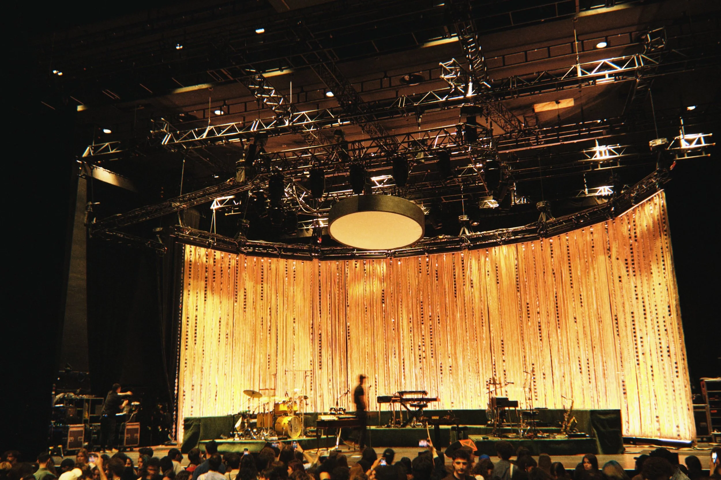 Stage setup with musical instruments and large golden curtain backdrop, audience seated awaiting event, overhead lighting and rigging equipment.