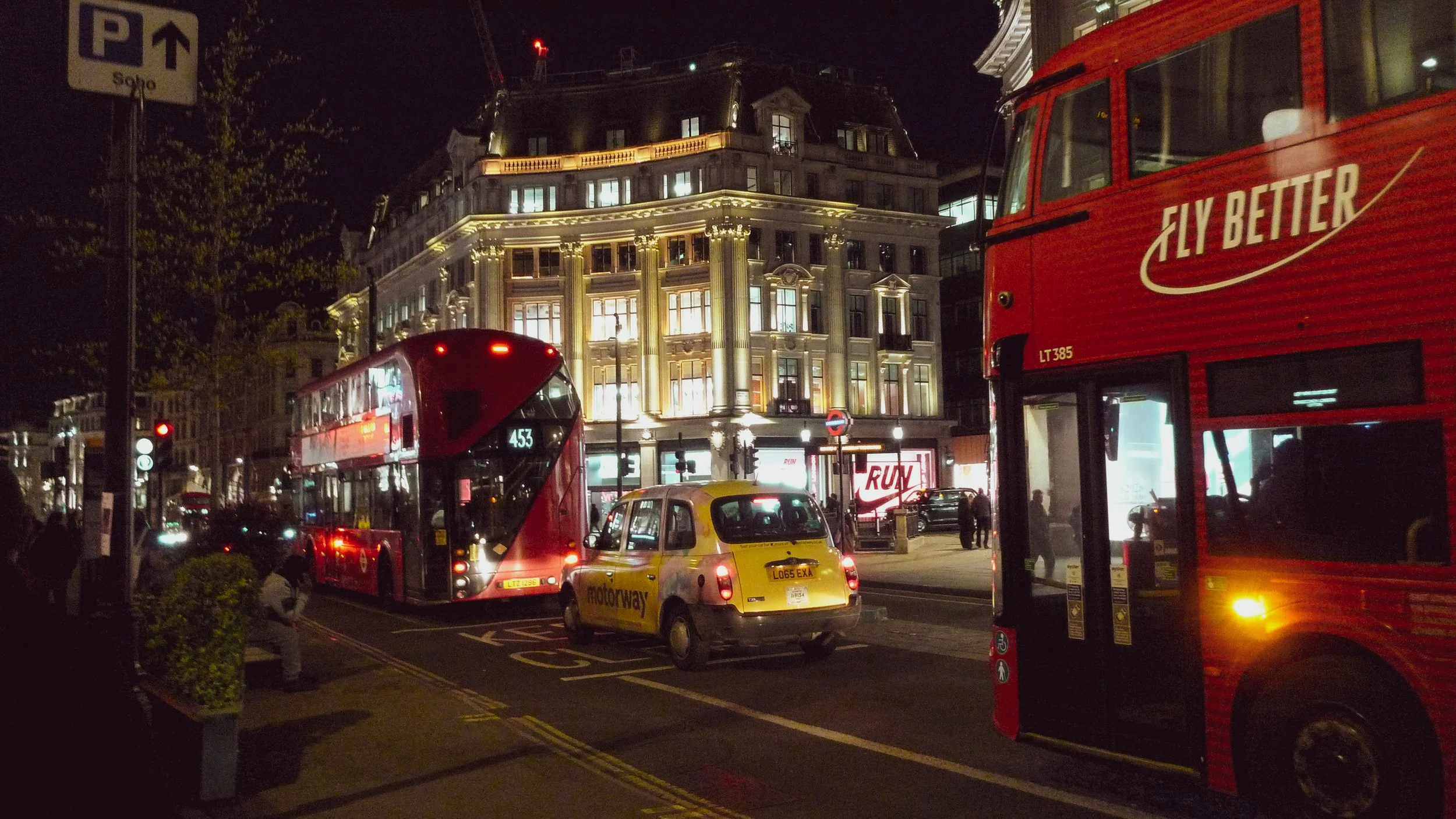 Nighttime city street scene with illuminated buildings, double-decker buses, taxis, and pedestrians.