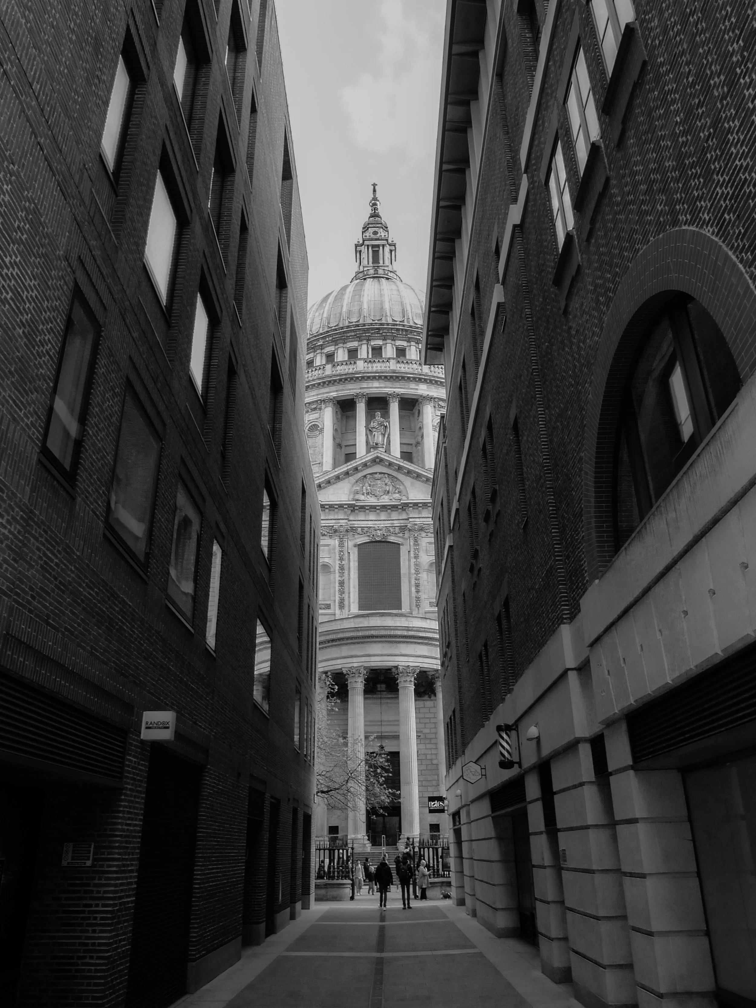 View through a narrow alleyway with brick buildings on both sides leading towards a distant domed building and a cathedral with a statue near the top of its entrance, overcast sky.