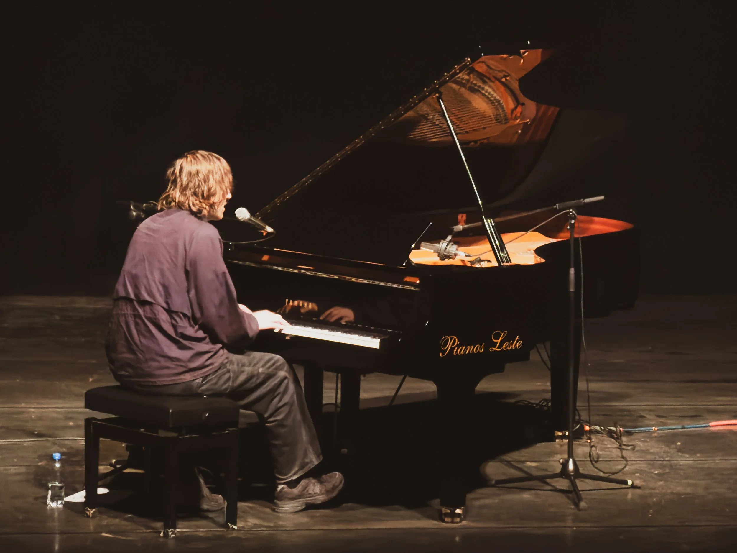 A man with long hair, wearing a purple shirt, sitting at a black grand piano on stage, playing music during a performance. The grand piano has the words 'Pianos Leste' on its side.