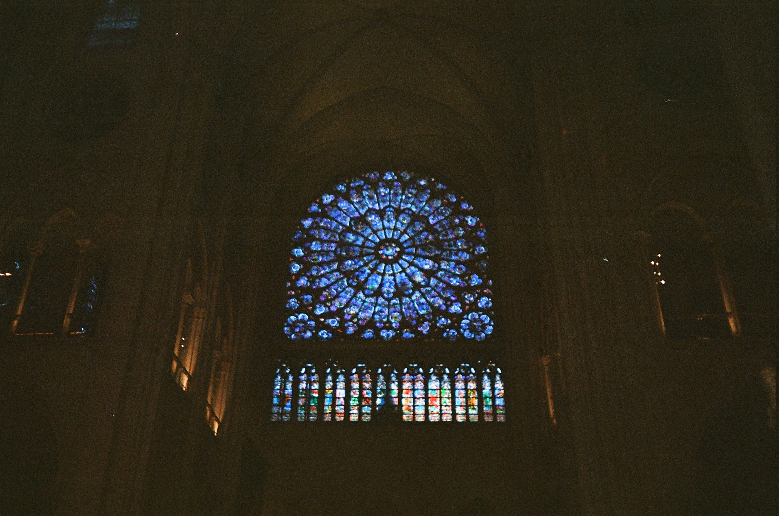 The interior of a cathedral showing a large, intricate stained glass rose window with vibrant colors, high arches, and Gothic architecture.