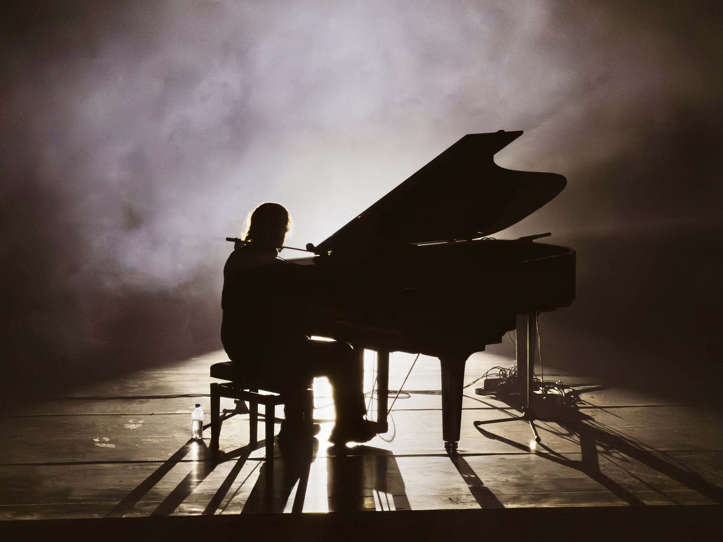 Silhouette of a person playing piano on stage, with a bright light behind them illuminating the foggy atmosphere.