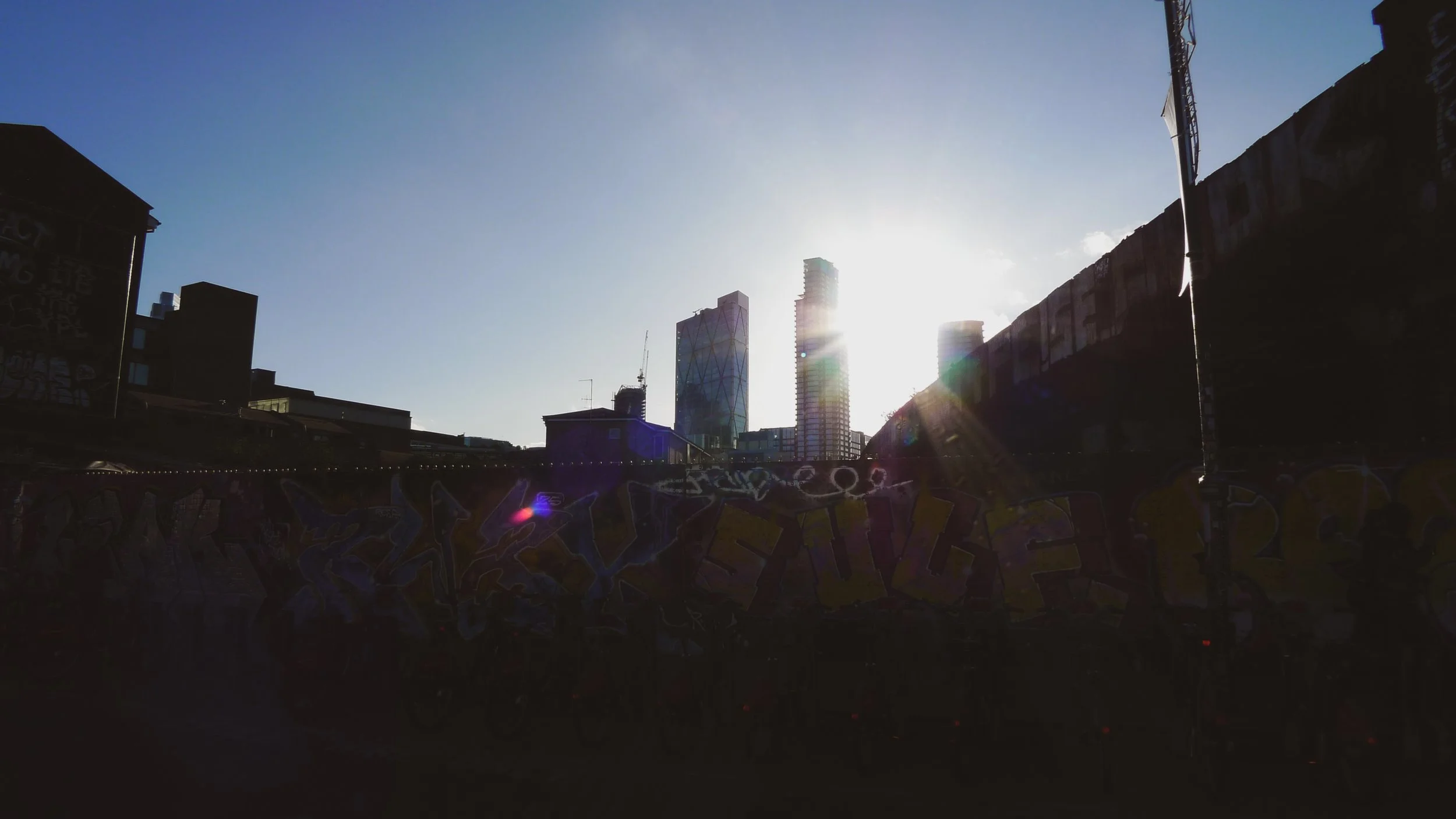 City skyline with tall modern buildings silhouetted against the bright sun, graffiti-covered wall in the foreground.