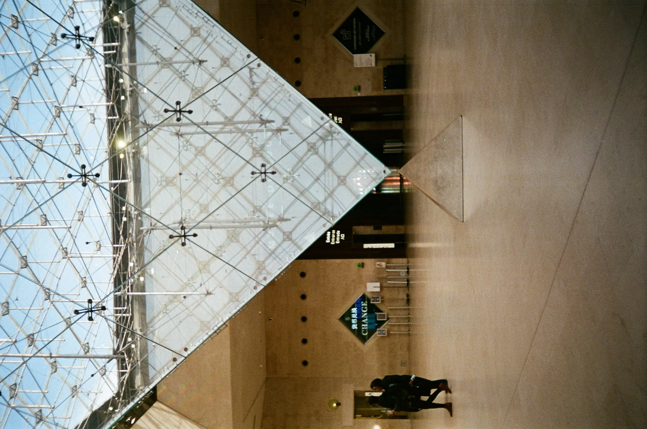 Interior of a museum or gallery featuring a large geometric sculpture or installation made of metal rods and crosses, displayed in front of a glass wall with a view of the sky. There are signs and people walking around in the background.