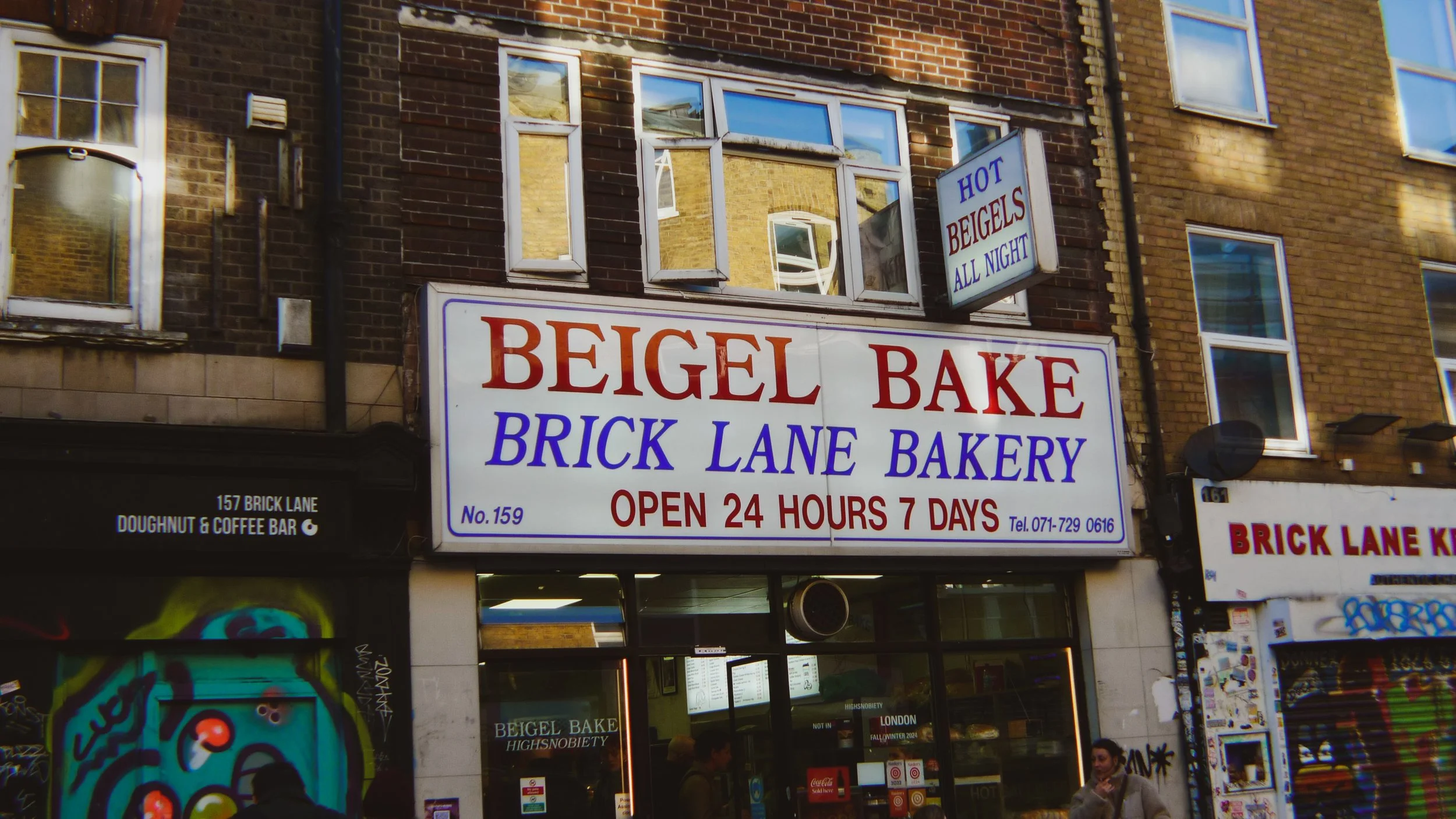 Sign for a bakery called 'Brick Lane Bake' offering hot beigels all night, located on Brick Lane in London, with open 24 hours, 7 days a week.
