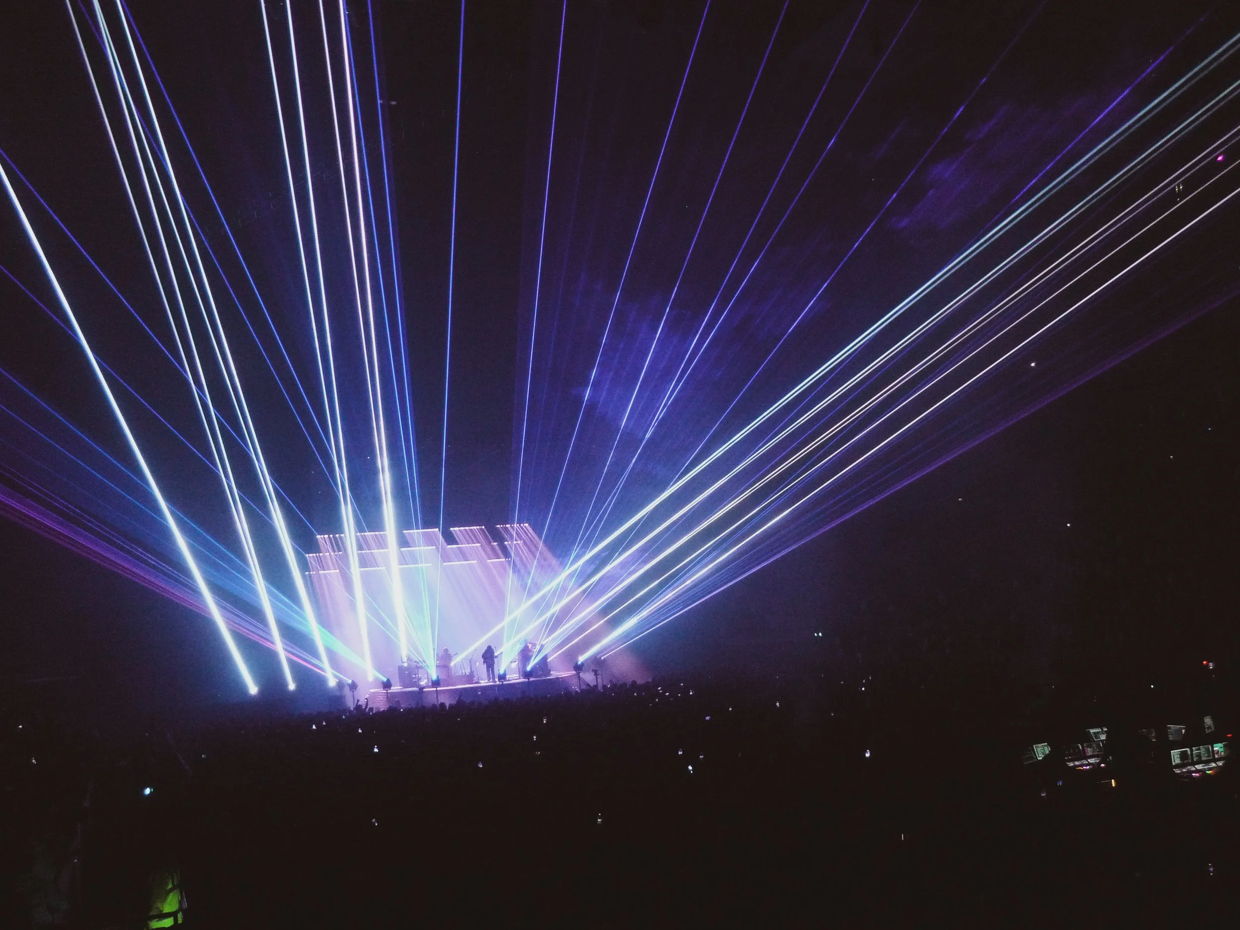 Nighttime scene of a concert or event with colorful laser lights projecting upward from the stage, illuminating the dark sky and silhouettes of the audience and performers.
