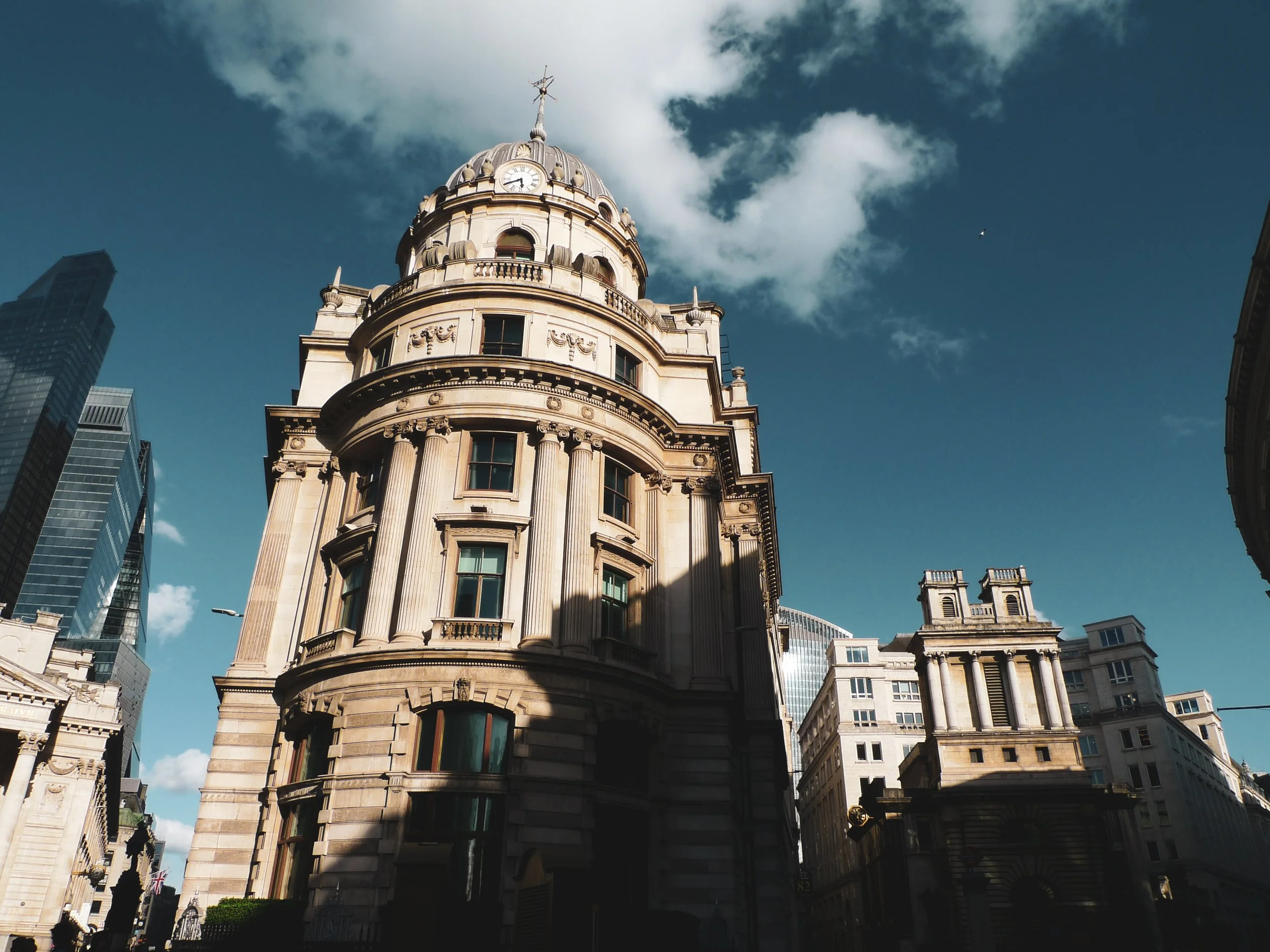 A historic building with a clock tower, ornate architectural details, and a weather vane on top, surrounded by modern skyscrapers against a blue sky with clouds.