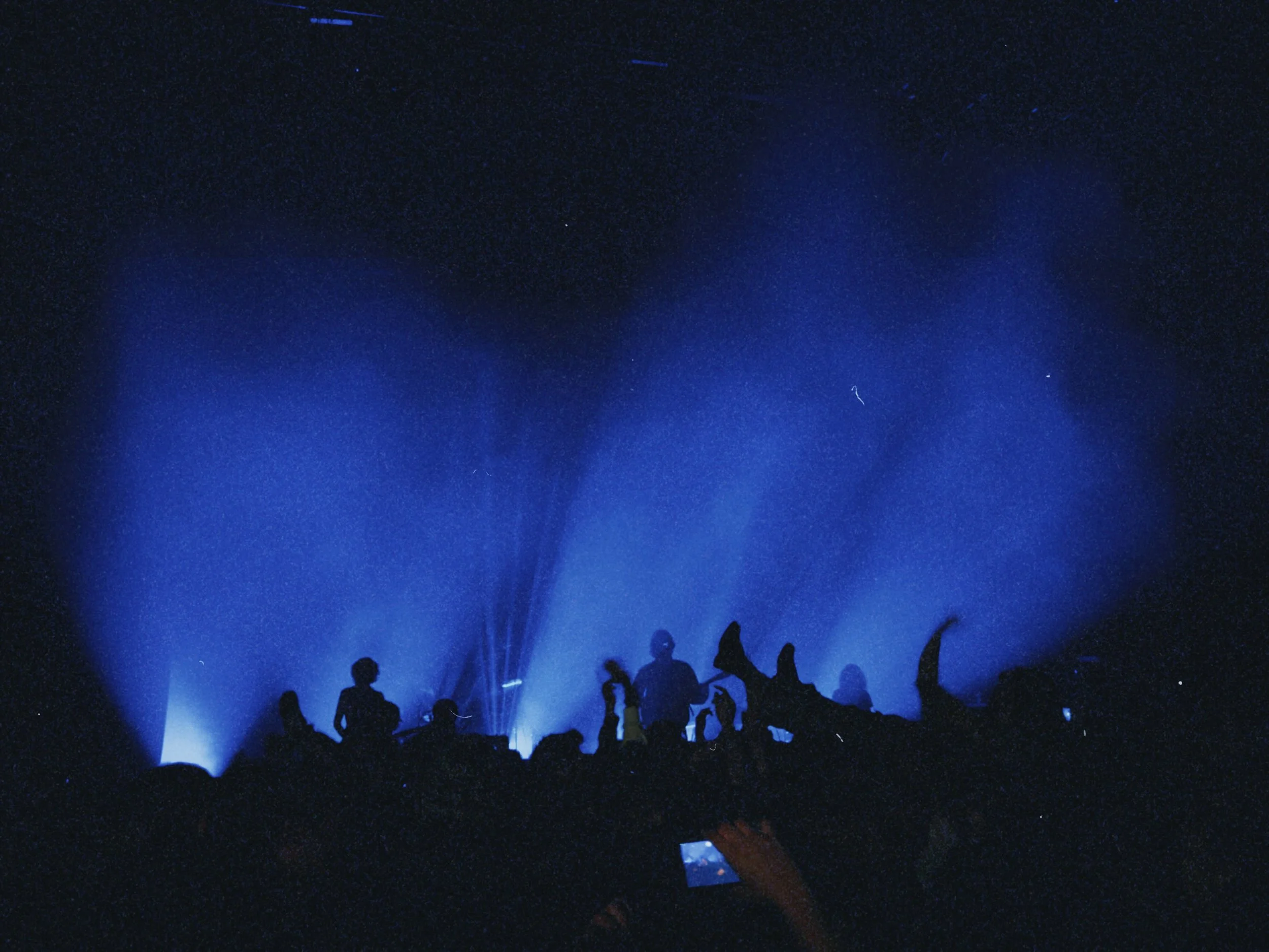 Dark blue night sky with bright stars and a faint faint streak of light. Silhouettes of people standing and sitting, possibly at an outdoor event or stargazing gathering, with some looking upwards at the sky.