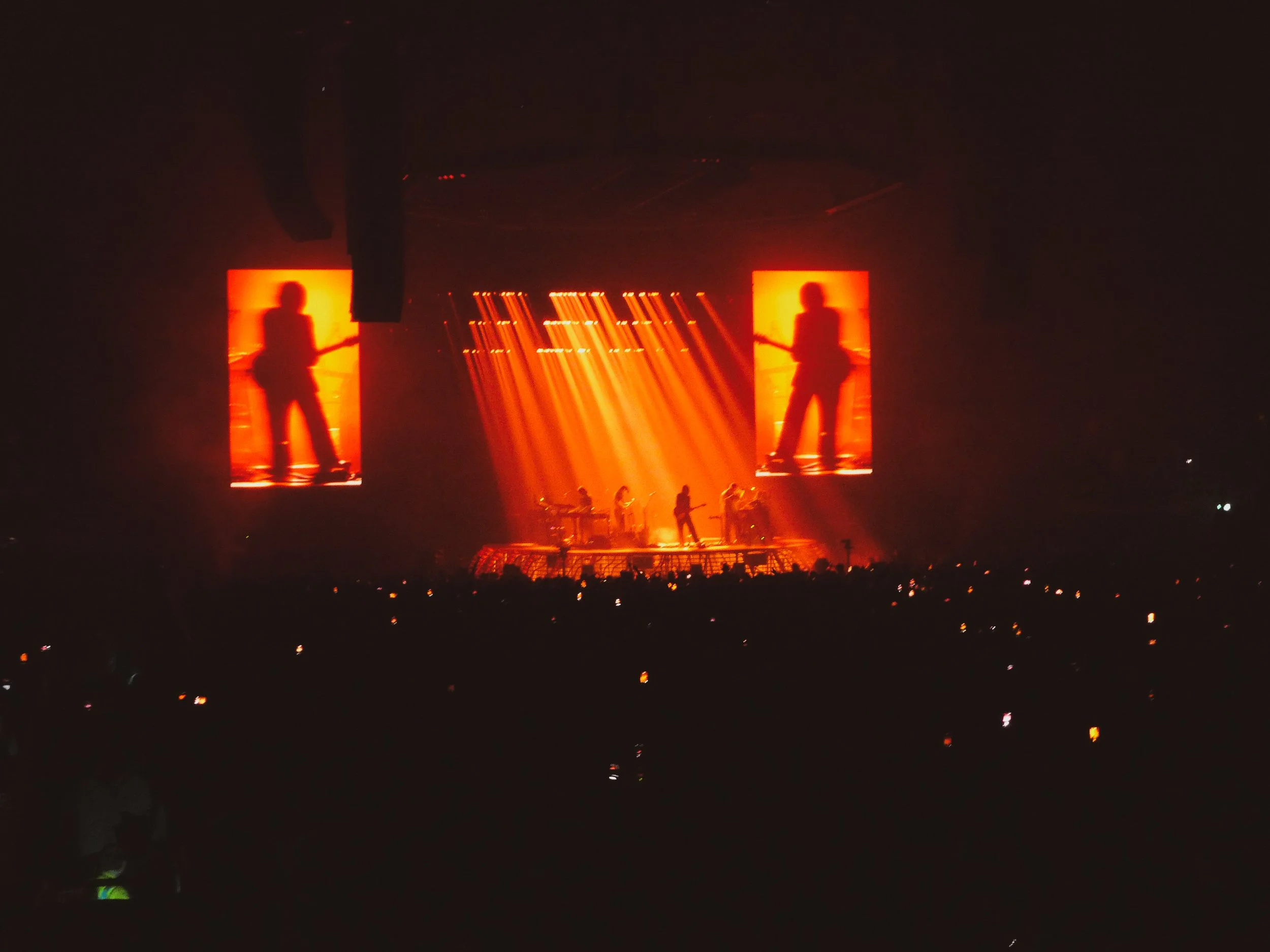 A concert stage with orange lighting, showing silhouettes of musicians and two large screens displaying the silhouette of a person playing guitar.