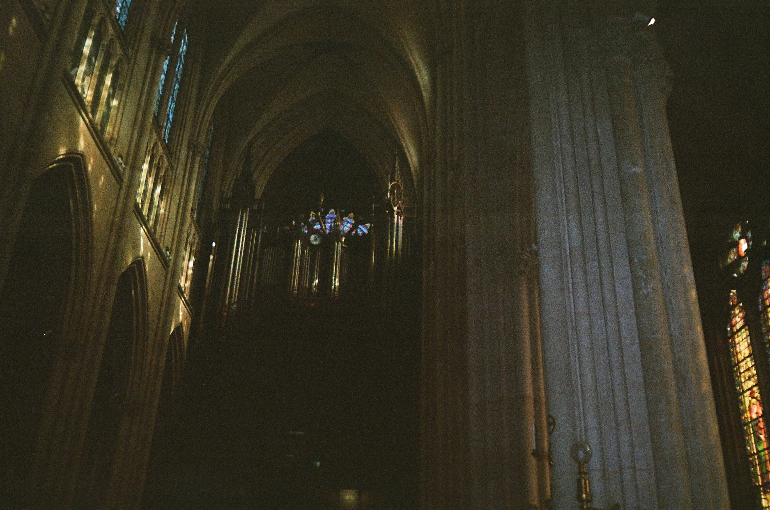 Interior view of a dimly lit cathedral with high vaulted ceilings, stained glass windows on the right, and illuminated chandeliers hanging from the ceiling.
