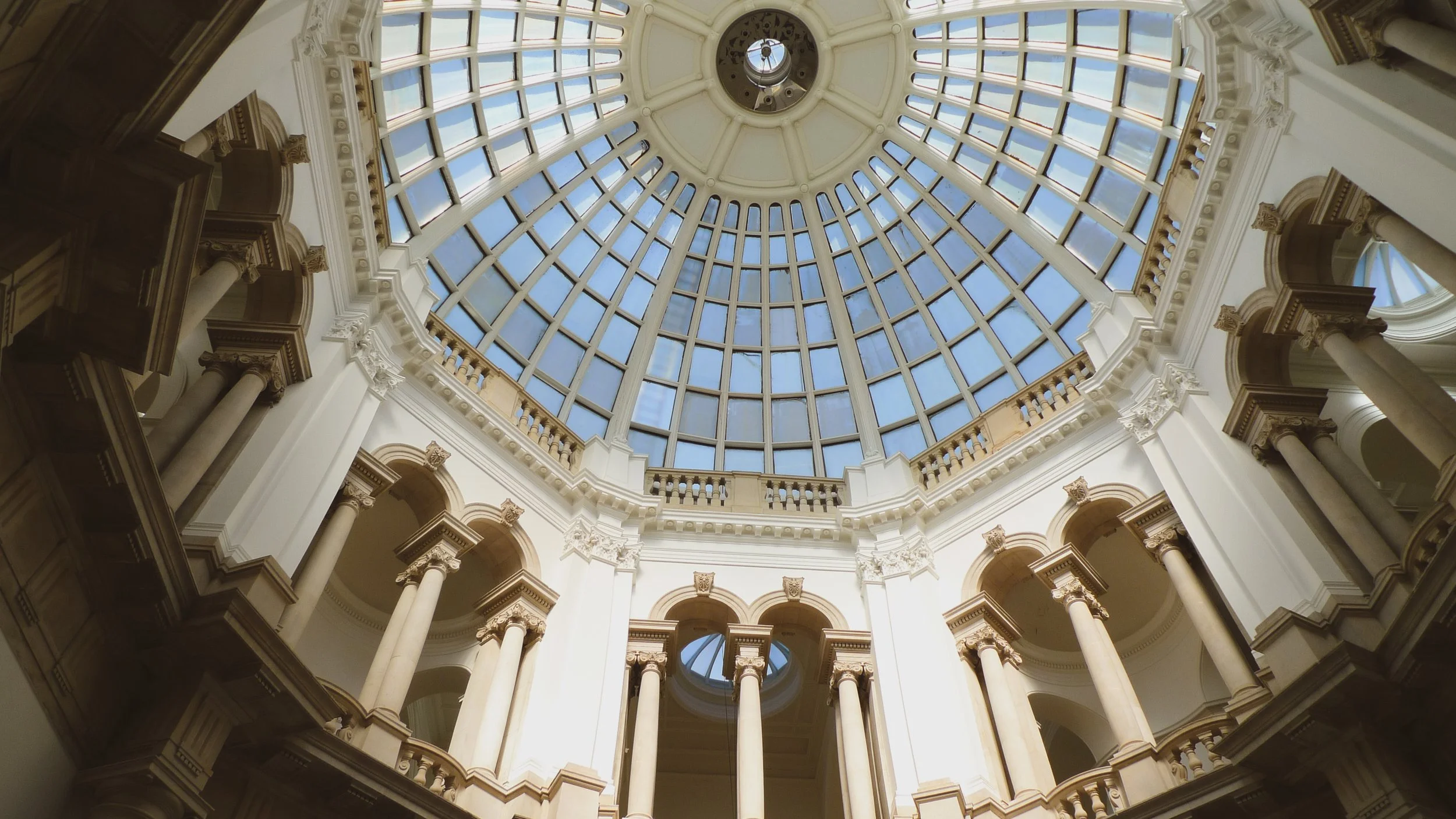 Interior view of a grand dome ceiling with a large glass skylight, ornate architectural details, columns, and arches.