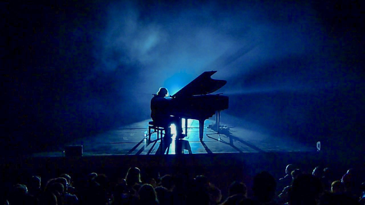 A person playing the piano on a stage illuminated with blue spotlight in front of an audience.