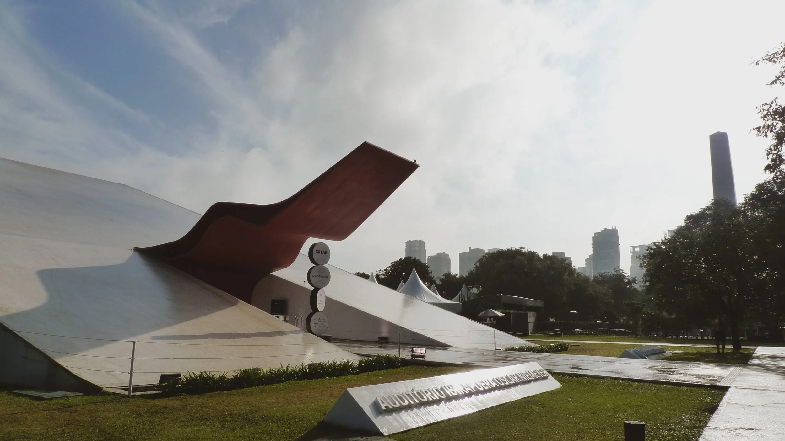 A modern sculpture with a curved, brown part and a white section underneath, located in an urban park with trees, tents, and a city skyline in the background.