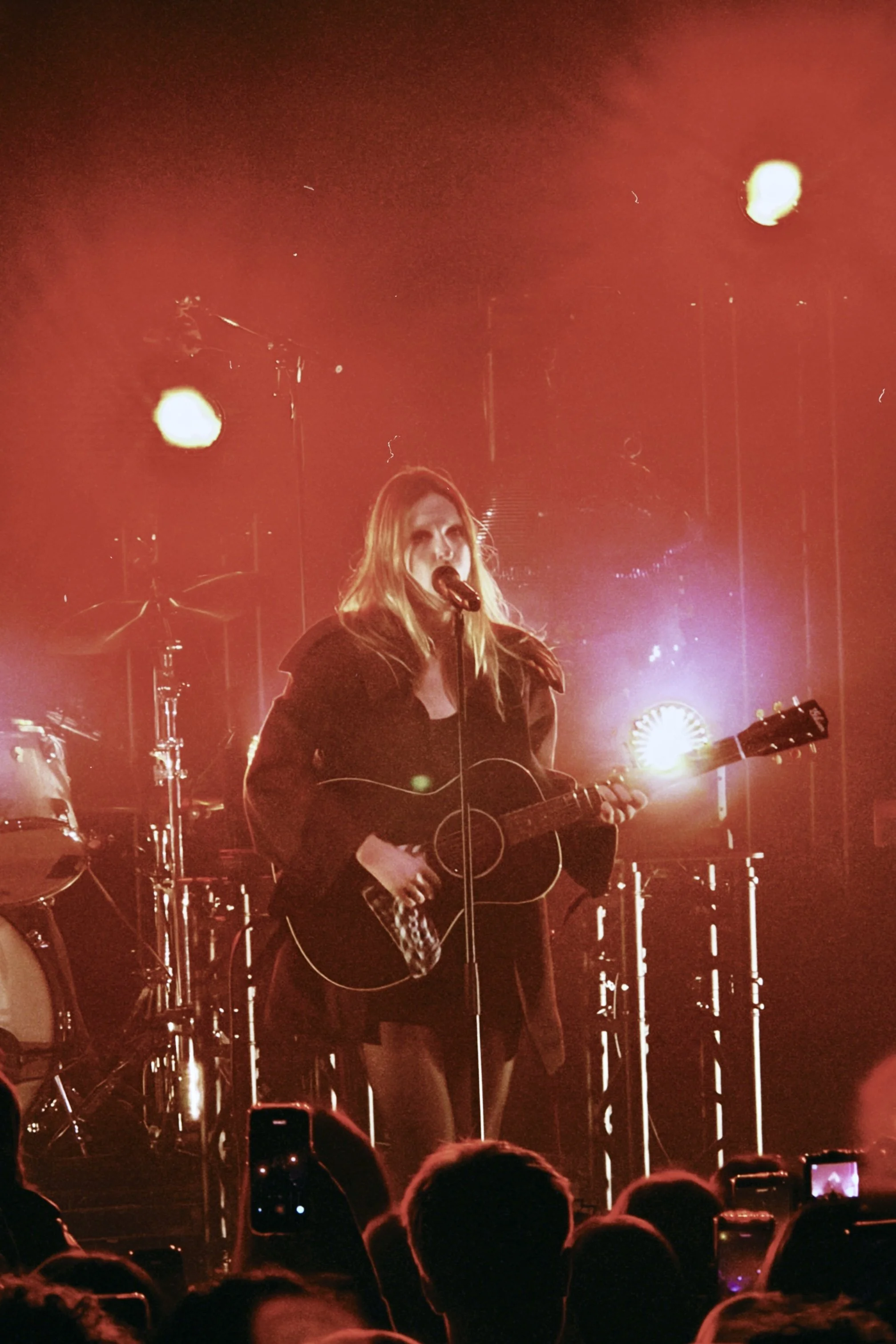 A woman with long hair and a dark outfit is performing on stage with an acoustic guitar and singing into a microphone, illuminated by red stage lighting and spotlights. The audience is visible in the foreground, taking photos.