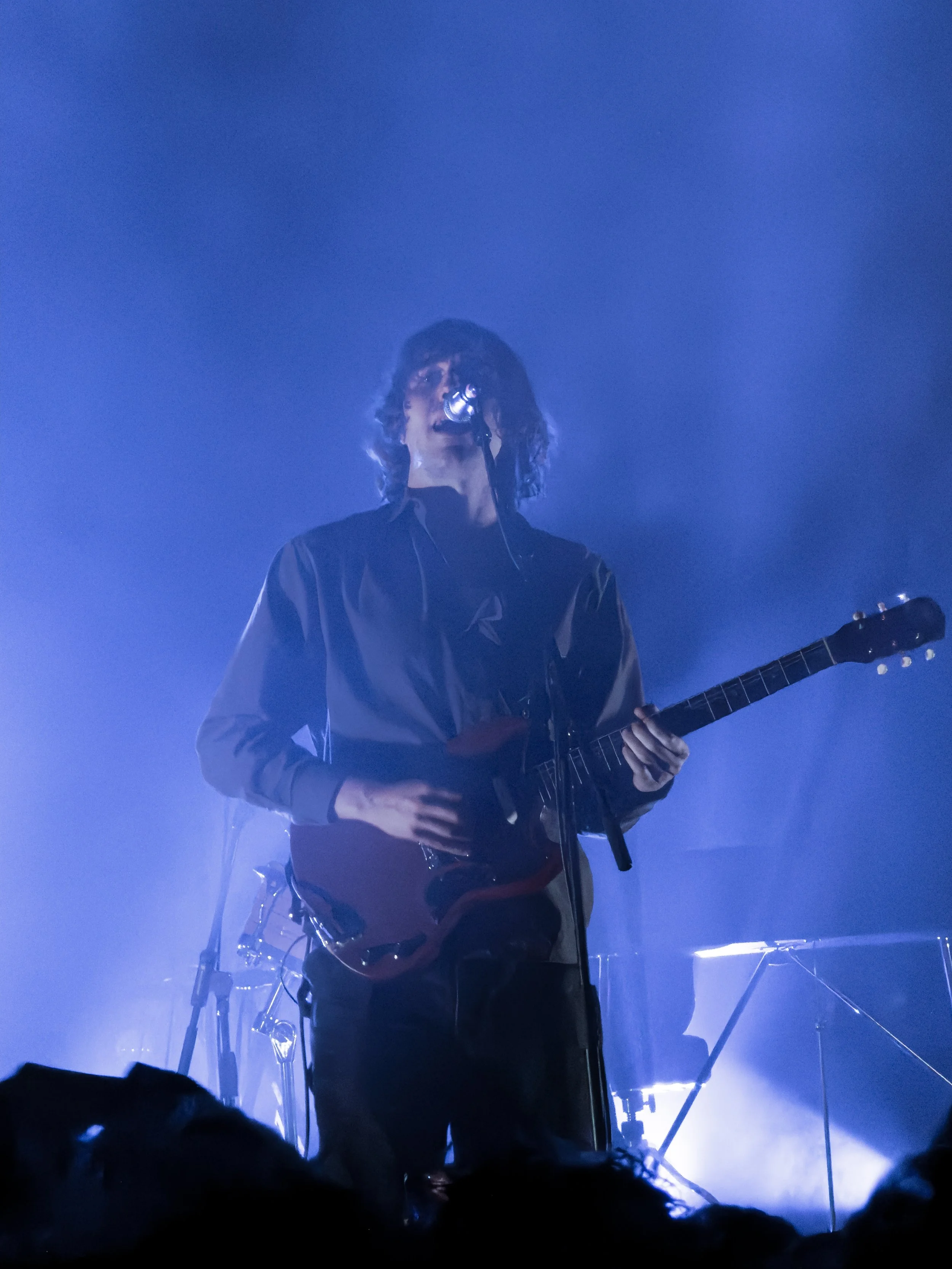 A musician with curly hair singing into a microphone while playing an electric guitar on stage with blue lighting.
