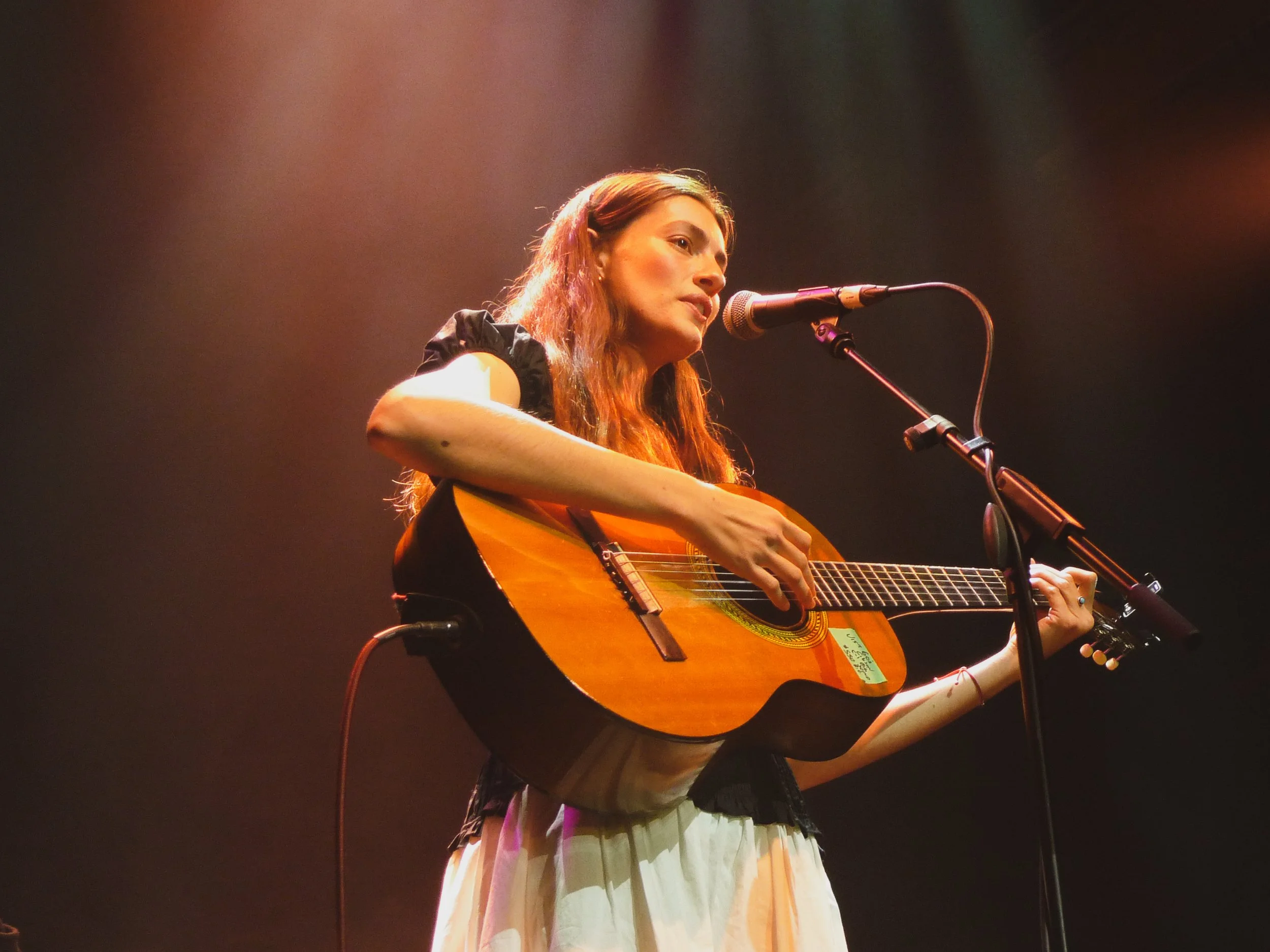 A woman with long brown hair singing into a microphone while playing an acoustic guitar on stage with a dark background.
